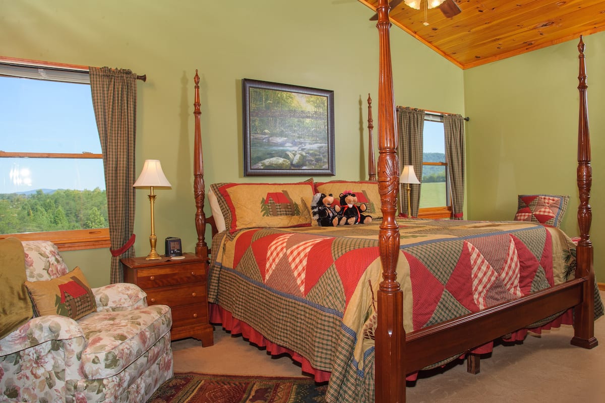 A four-poster bed with a quilt, a nightstand with a lamp, a floral armchair, and a large window with a view of greenery.