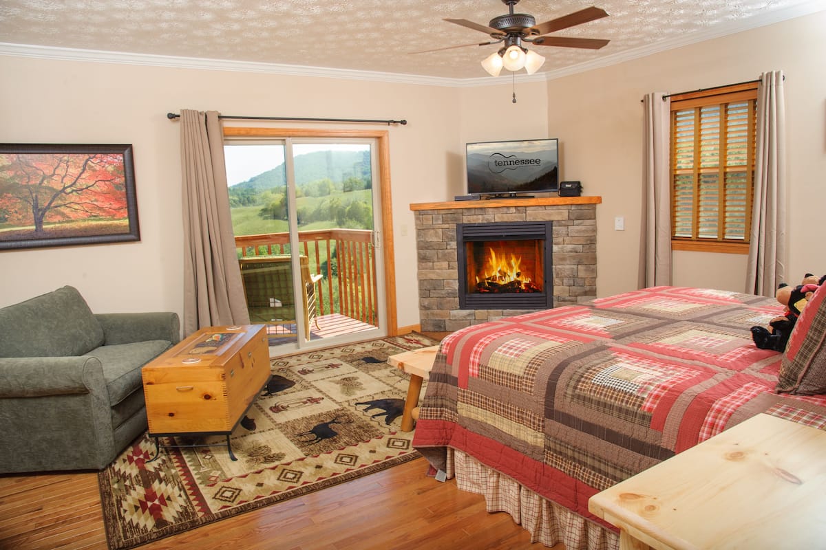 A bedroom with a fireplace, a sofa, a wooden chest, a bed with a quilt, a television, and a balcony with a view of the outdoors.