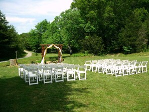Preparación para una ceremonia de boda al aire libre, con sillas blancas dispuestas en filas y un arco de madera decorado con flores.