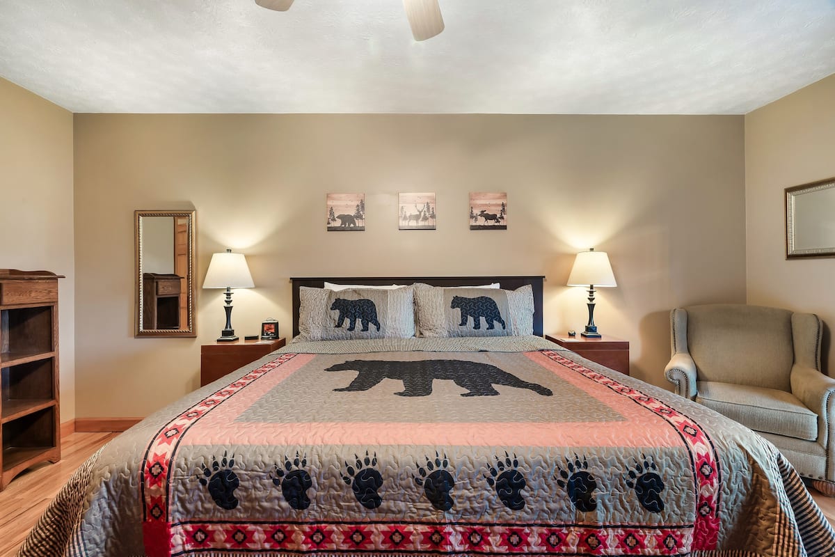 A bedroom with a large bed, two bedside lamps, a chair, a mirror, and three wall-mounted pictures.