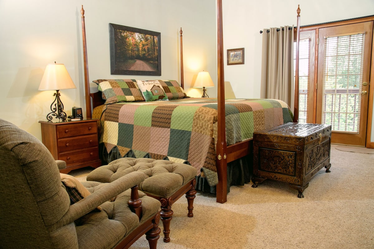 A four-poster bed with a quilt, two bedside lamps, a wooden nightstand, a tufted armchair, and a small wooden chest of drawers.