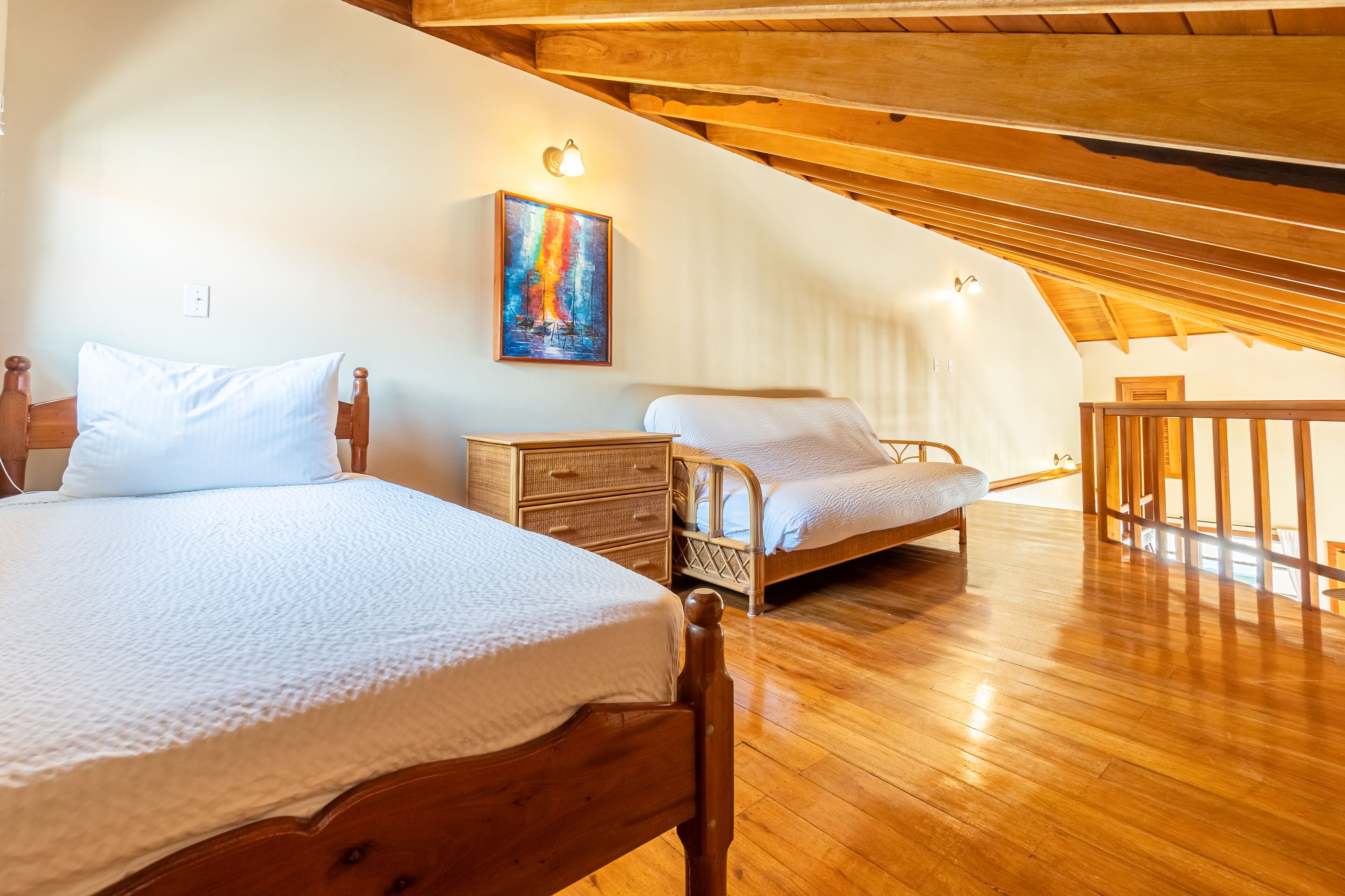 A bedroom with two beds, wooden flooring, a painting on the wall, and a sloped ceiling.
