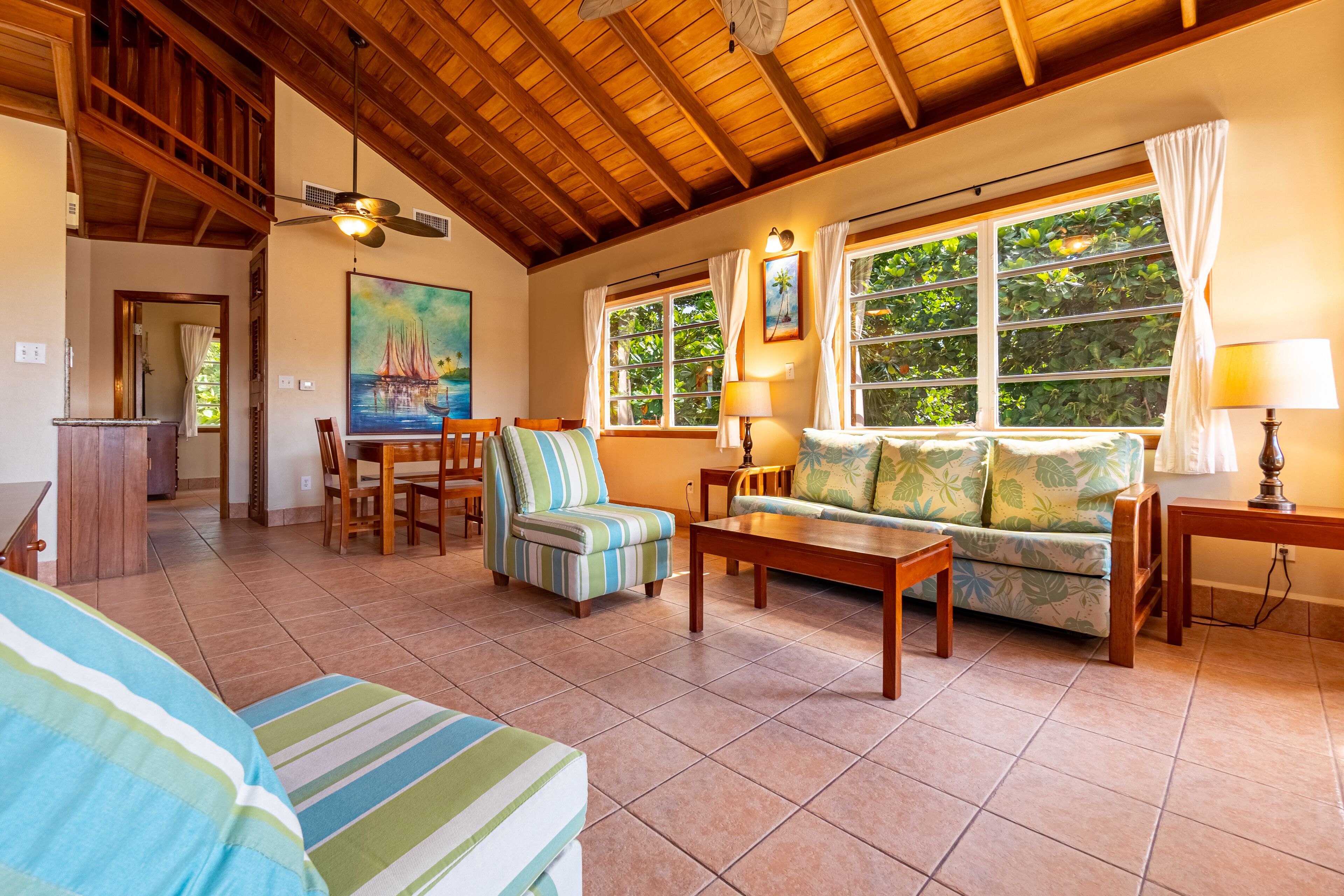 A spacious living area with a high ceiling, wooden beams, a dining set, a sofa with patterned cushions, and large windows offering a view of greenery.