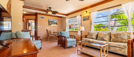 A living room with a wooden coffee table, blue armchairs, a sofa with tropical print, and a ceiling fan.
