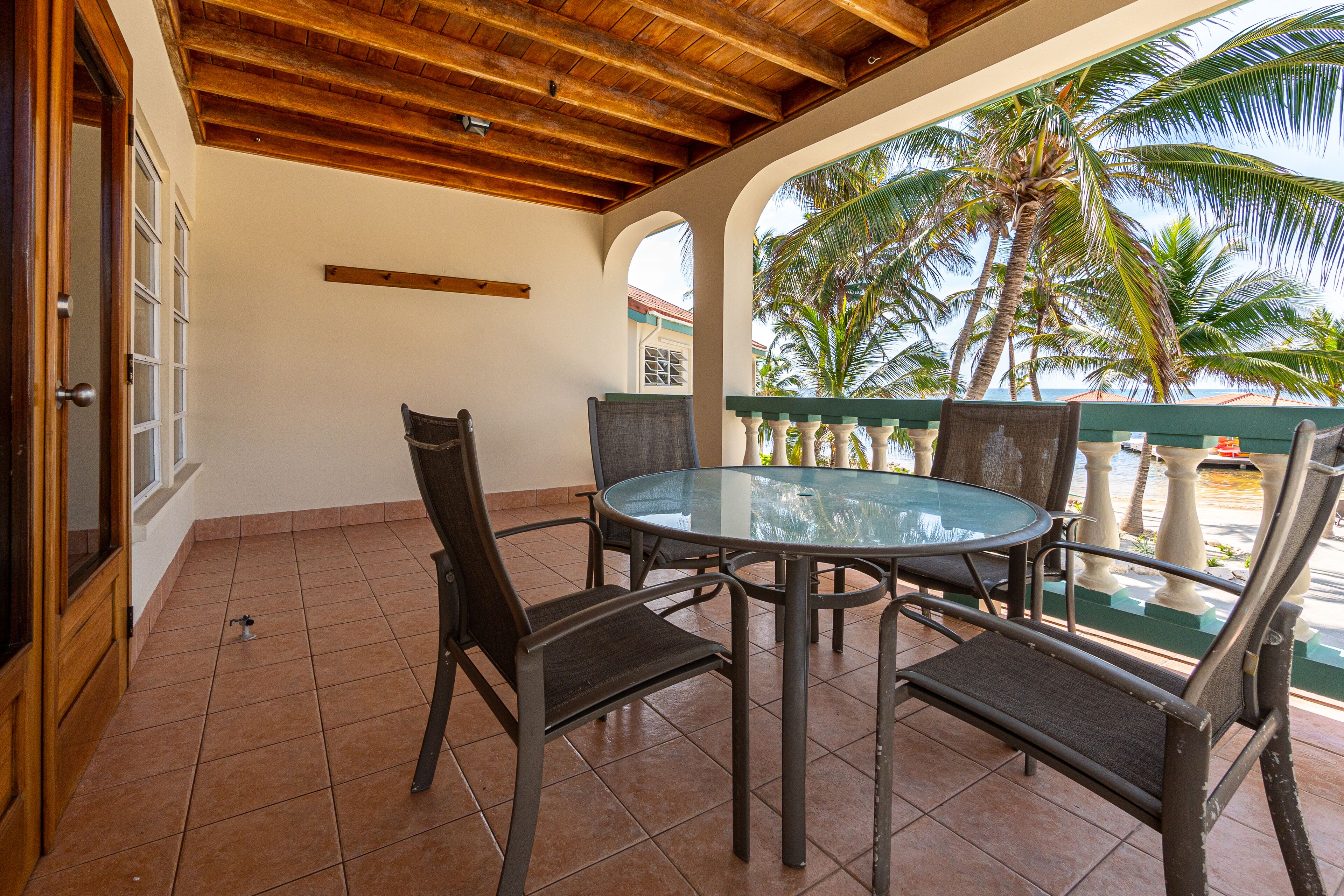Une terrasse couverte avec une table ronde en verre et quatre chaises, un plafond en bois et une vue sur les palmiers.