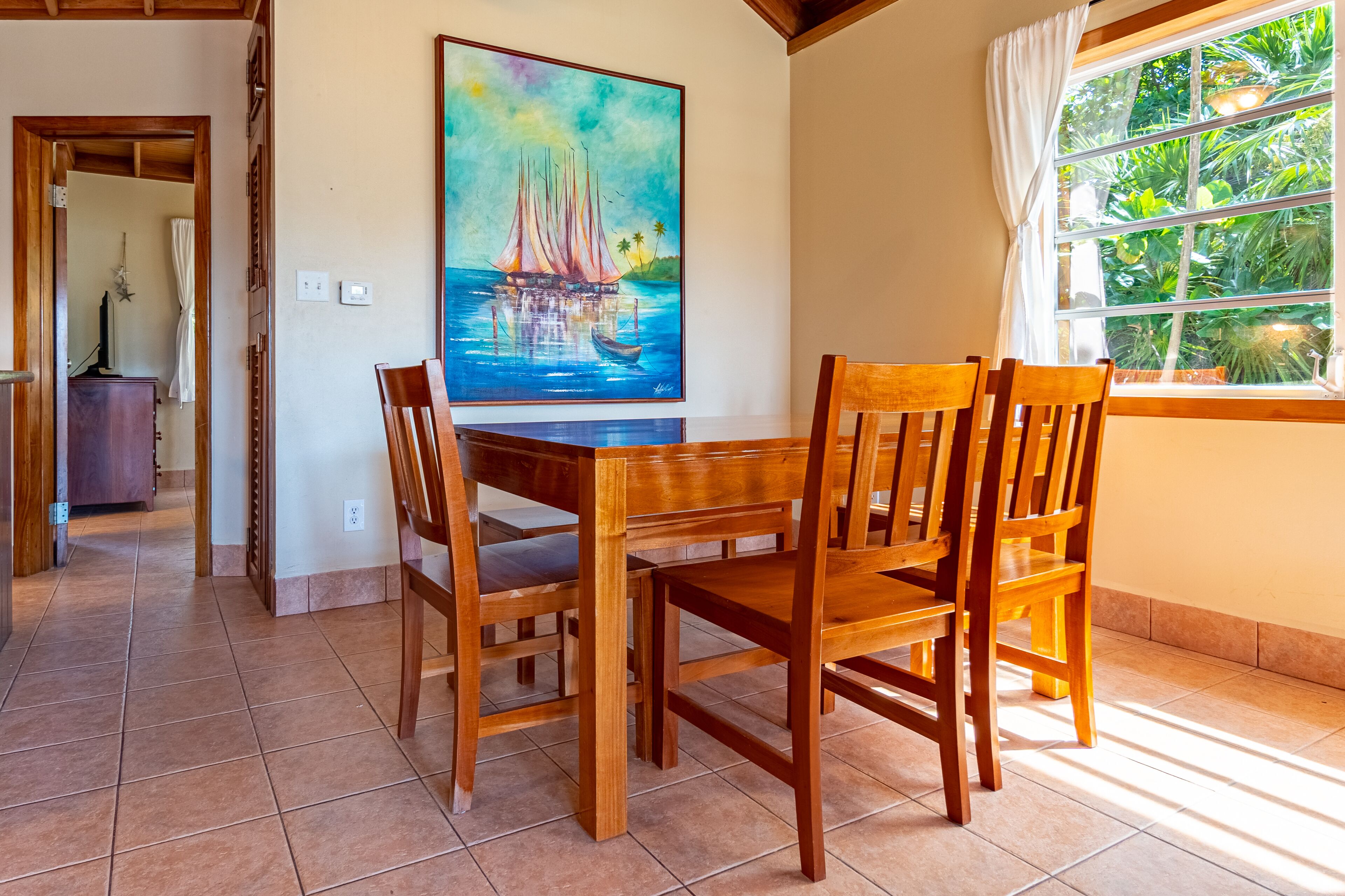Un coin repas avec des meubles en bois, un tableau représentant un voilier et une vue sur la verdure par la fenêtre.