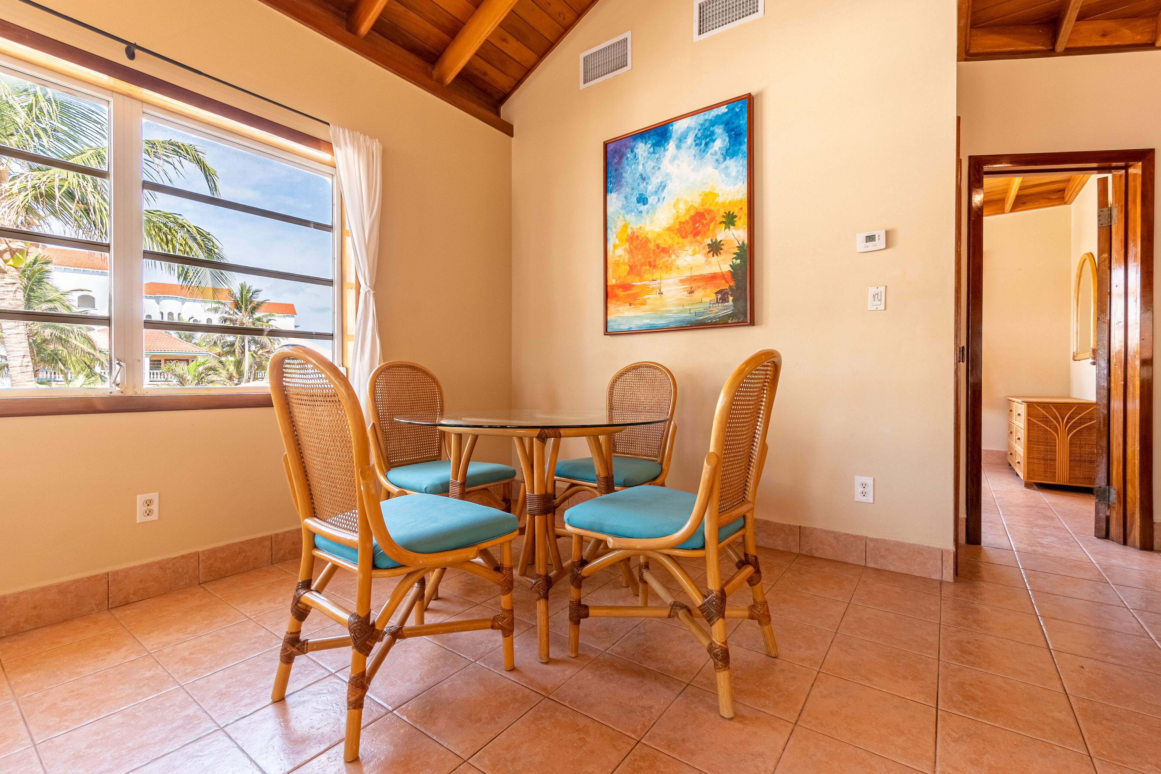 A dining area with wicker chairs and a round table, a painting of a sunset, and a view of palm trees through a window.