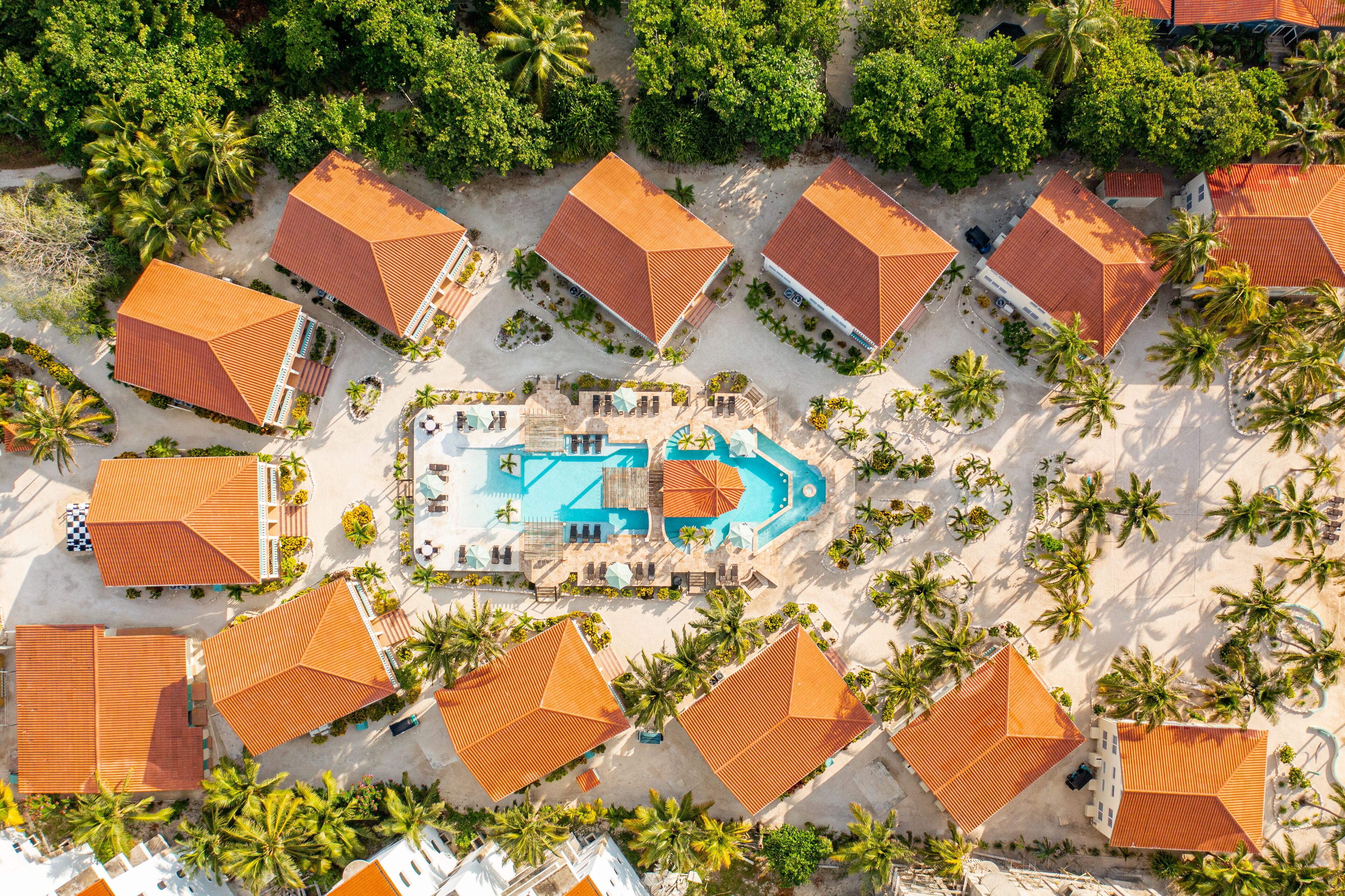 An aerial view of a resort with multiple buildings, a central swimming pool, and surrounding palm trees.
