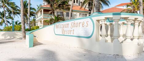 A sign for Belizean Shores Resort with a white and teal color scheme, surrounded by palm trees and a building with a red roof.