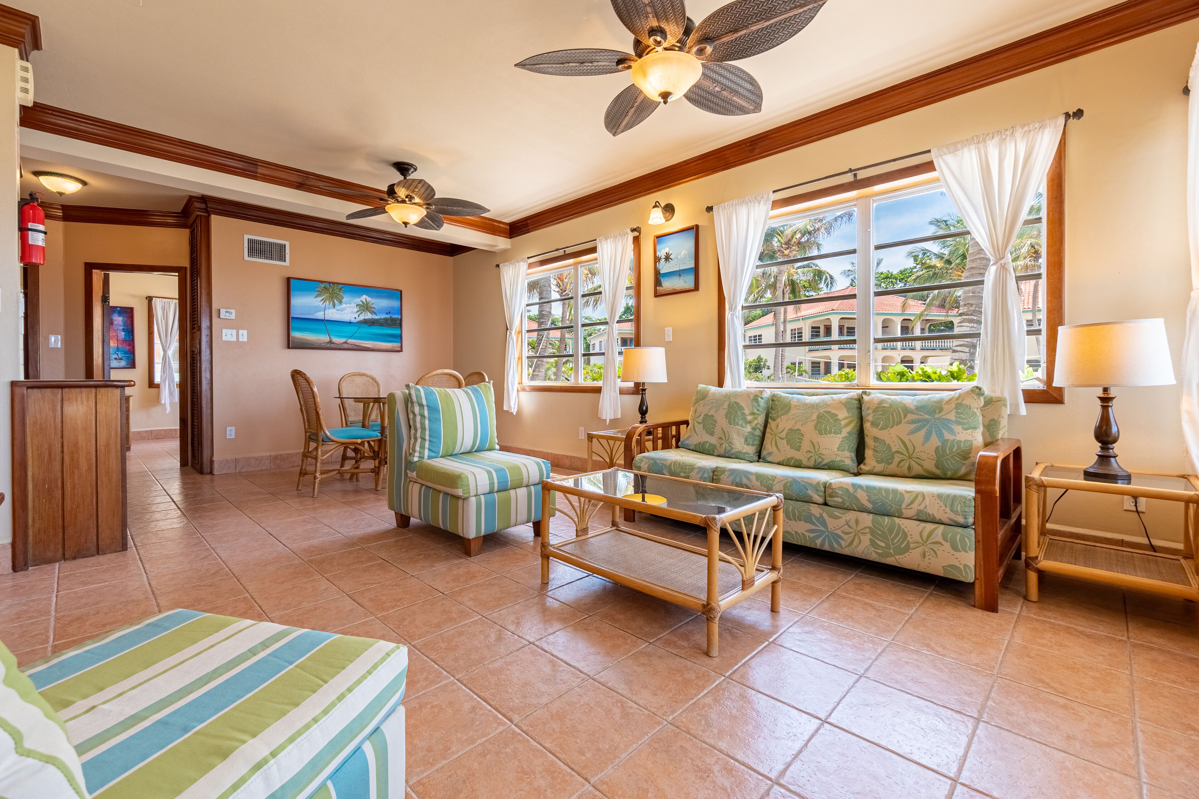 A spacious living room with a large sofa, a coffee table, and a ceiling fan. There are windows with curtains, a painting, and a view of palm trees.