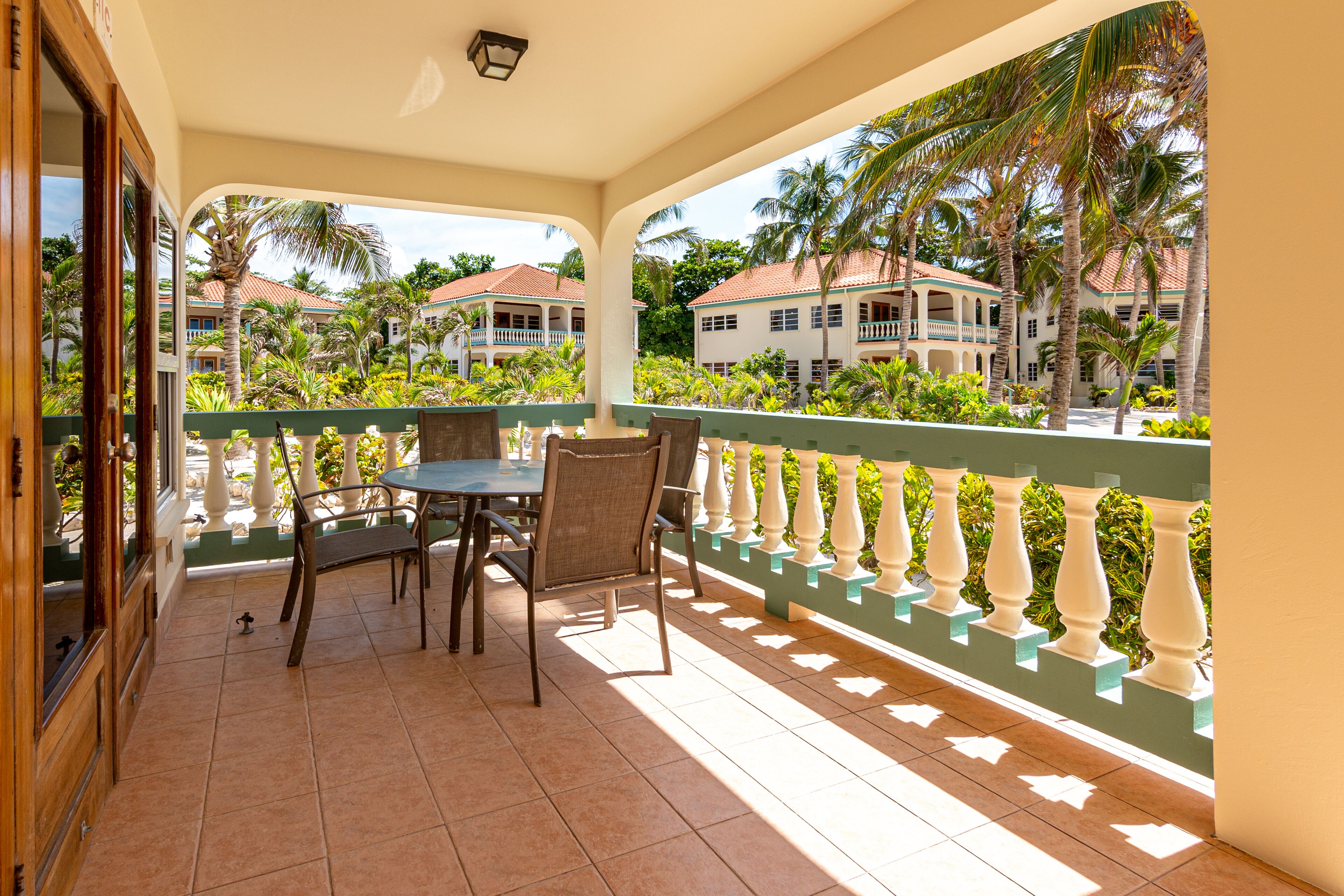 A covered balcony with a table and chairs, overlooking a lush garden and buildings.
