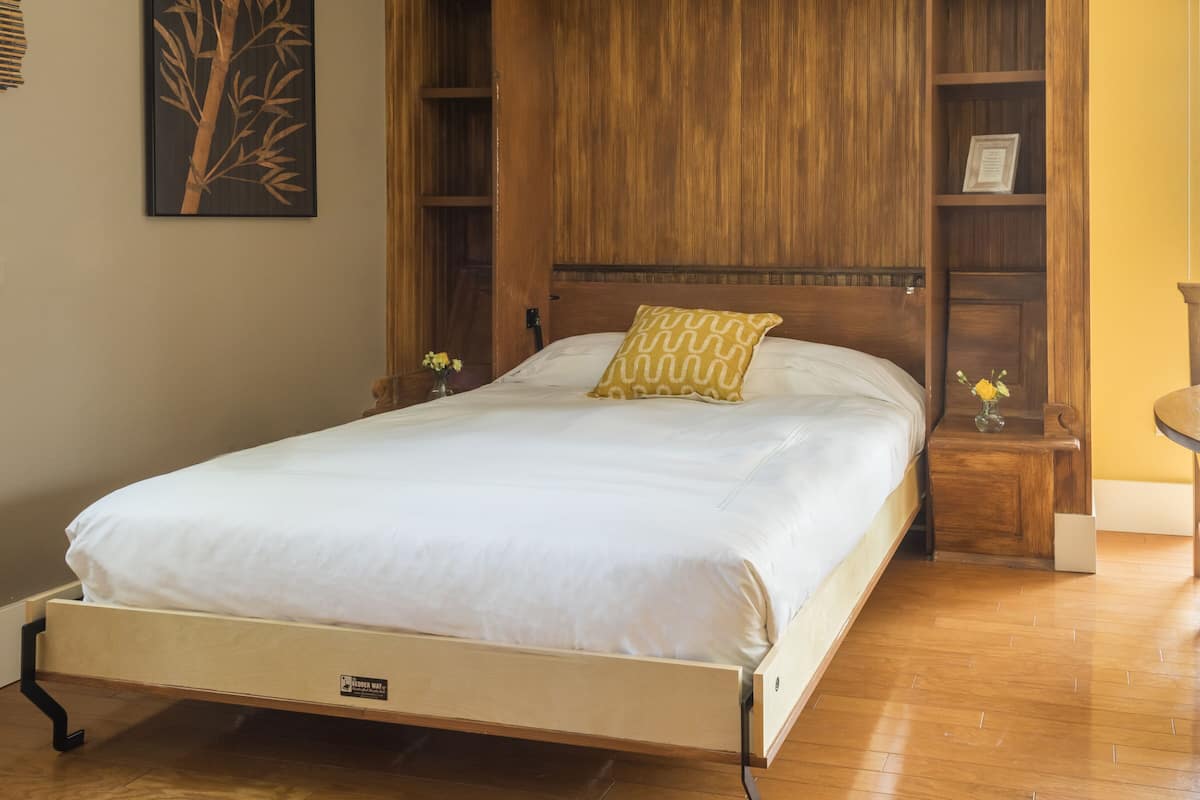 A bedroom with a wooden bed frame, a white mattress, and a yellow patterned pillow. There are wooden nightstands with flower vases on them.