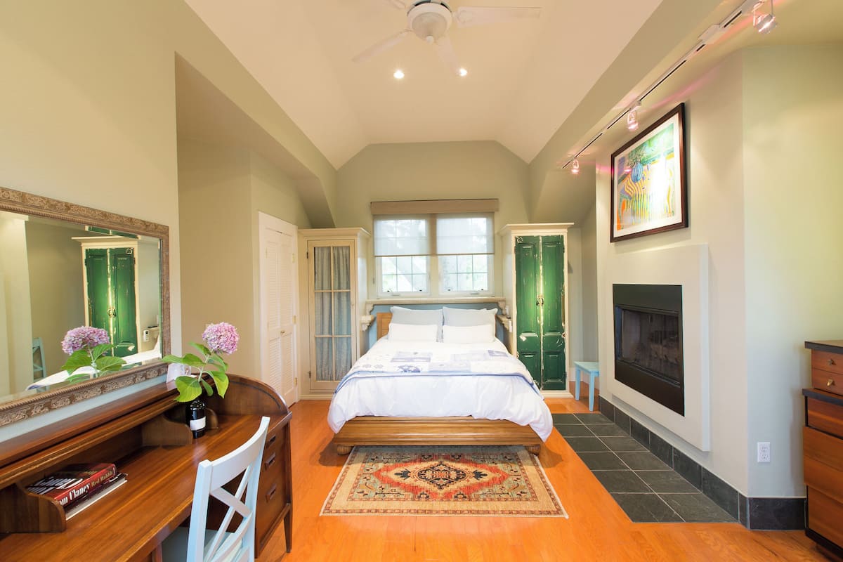 A bedroom with a bed, a desk, a chair, a mirror, a fireplace, and a window.