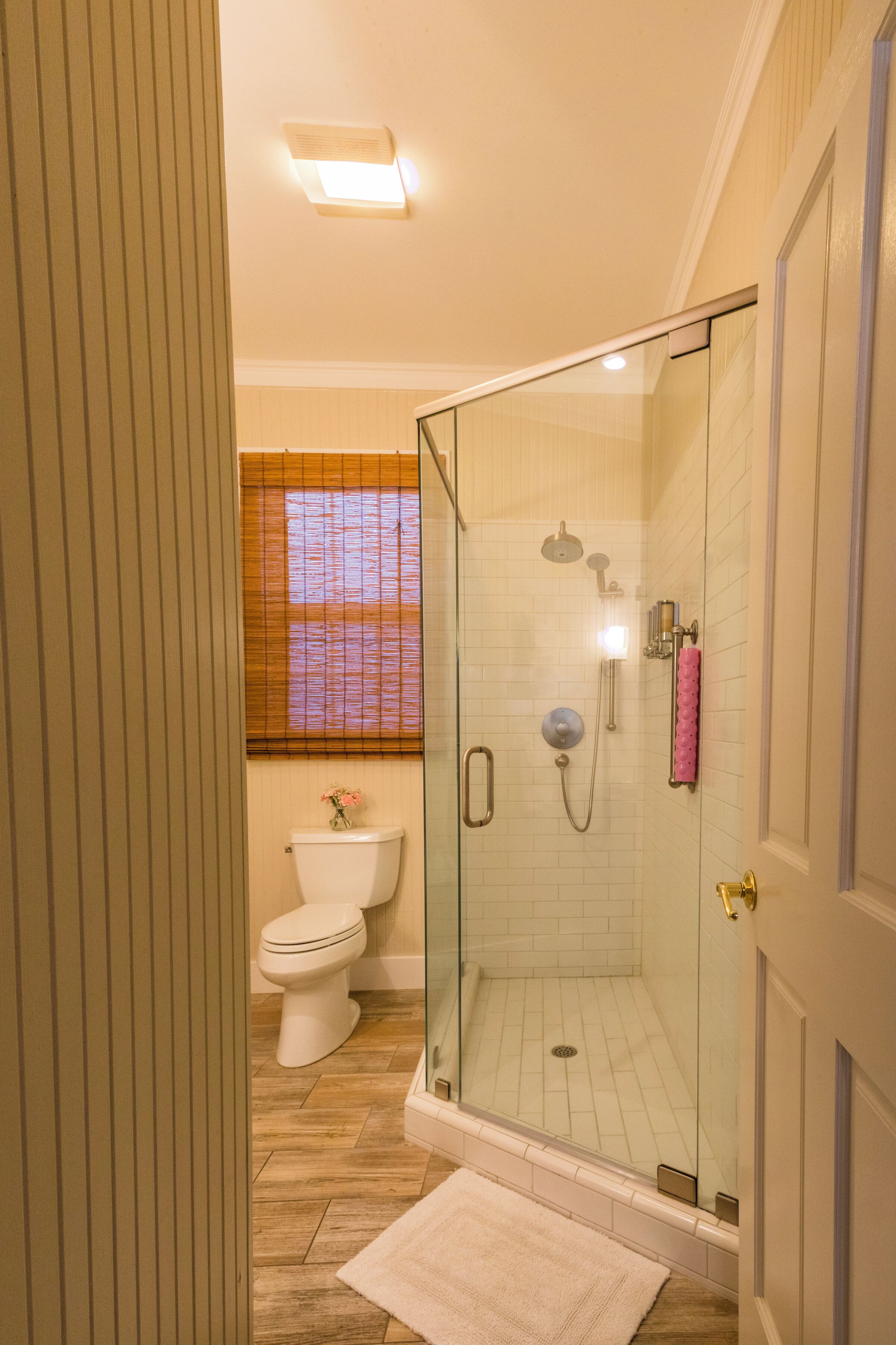 A glass-enclosed shower with a handheld showerhead, a toilet, and a window with blinds.