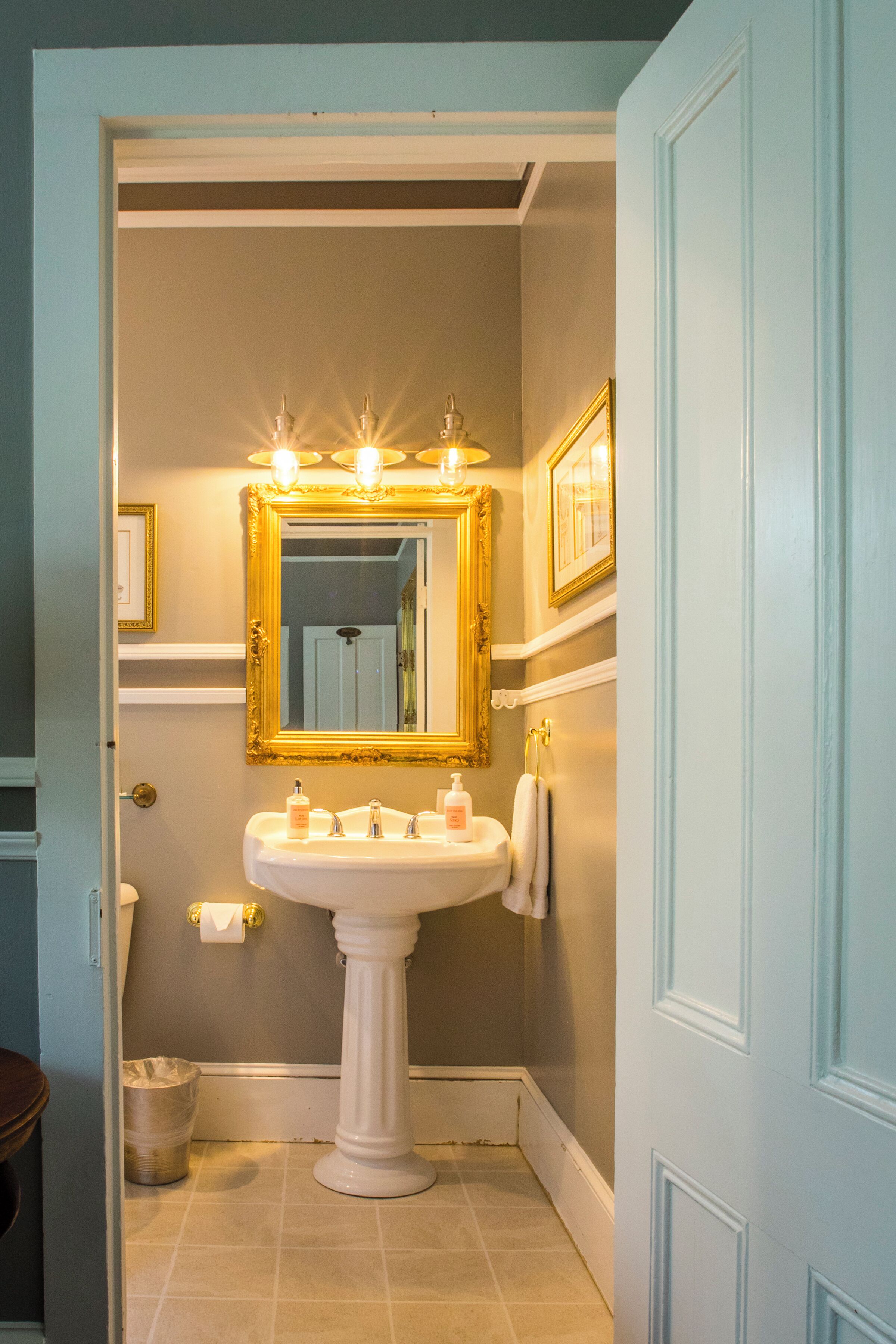 Ein Badezimmer mit einem Standwaschbecken, einem großen Spiegel mit goldener Rahmung und einer Leuchte darüber.