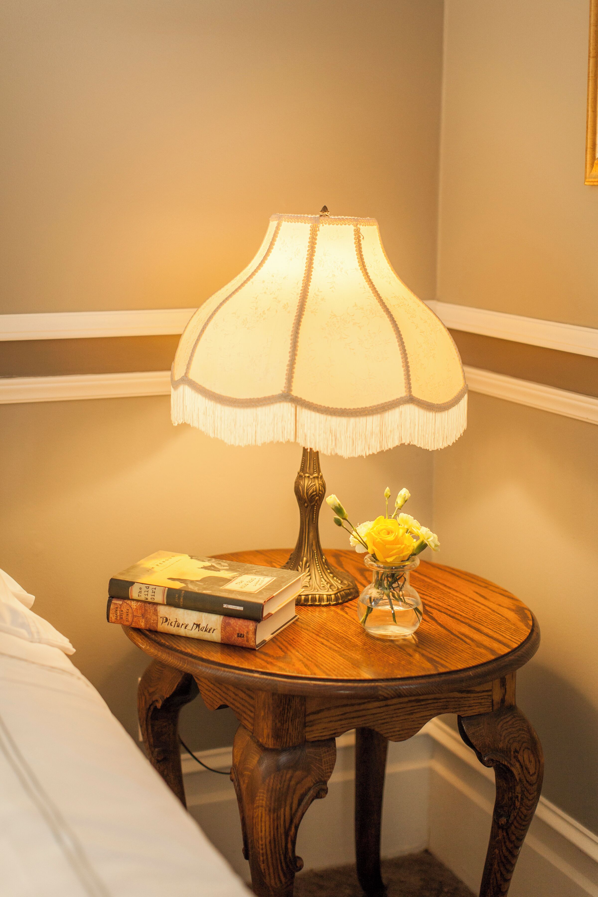 A wooden bedside table with a lamp, a vase with flowers, and a stack of books.
