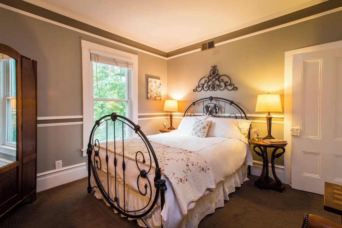 A bedroom with a four-poster bed, two bedside lamps, a window with blinds, and a door.