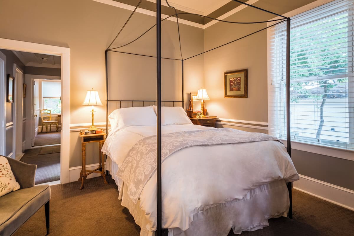 A four-poster bed with a canopy, bedside tables with lamps, a chair, and a window with blinds.