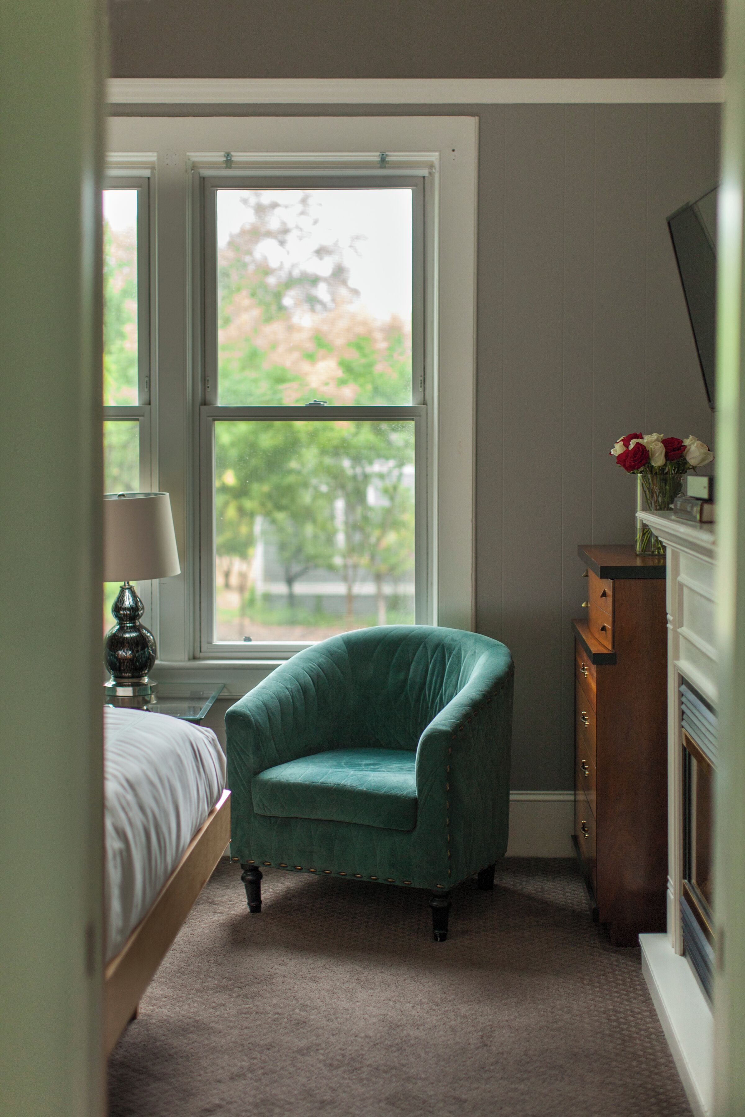 A bedroom with a bed, a teal armchair, a dresser, and a window with a view of trees.