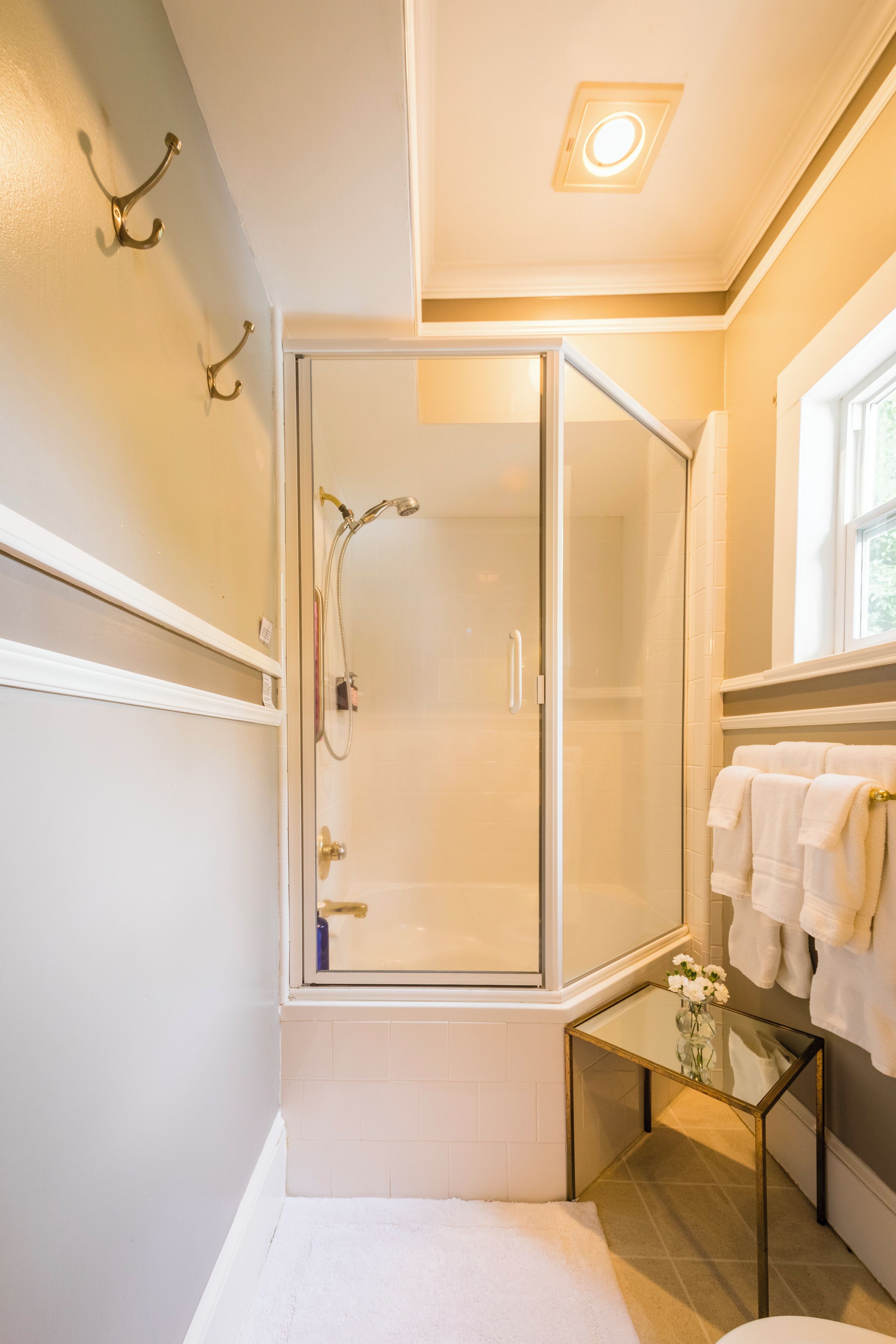 A bathroom with a glass shower enclosure, a small table with a vase, and a window with towels.