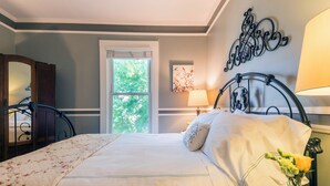 A bedroom with a bed, a bedside table with a lamp, a window with a view of trees, and a decorative wall piece. - Inn St Helena (St. Helena)