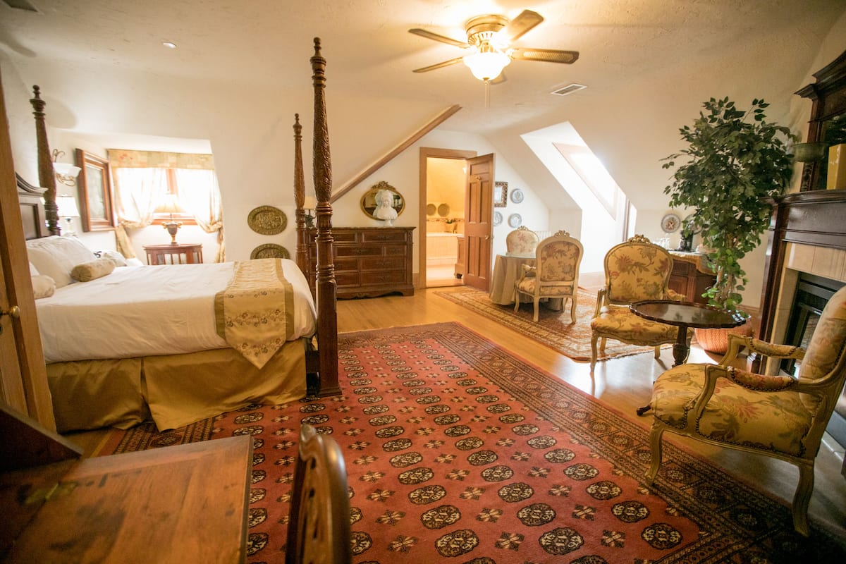 A bedroom with a four-poster bed, a wooden dresser, a red patterned rug, a chandelier, and a view of the dining area through an open door.