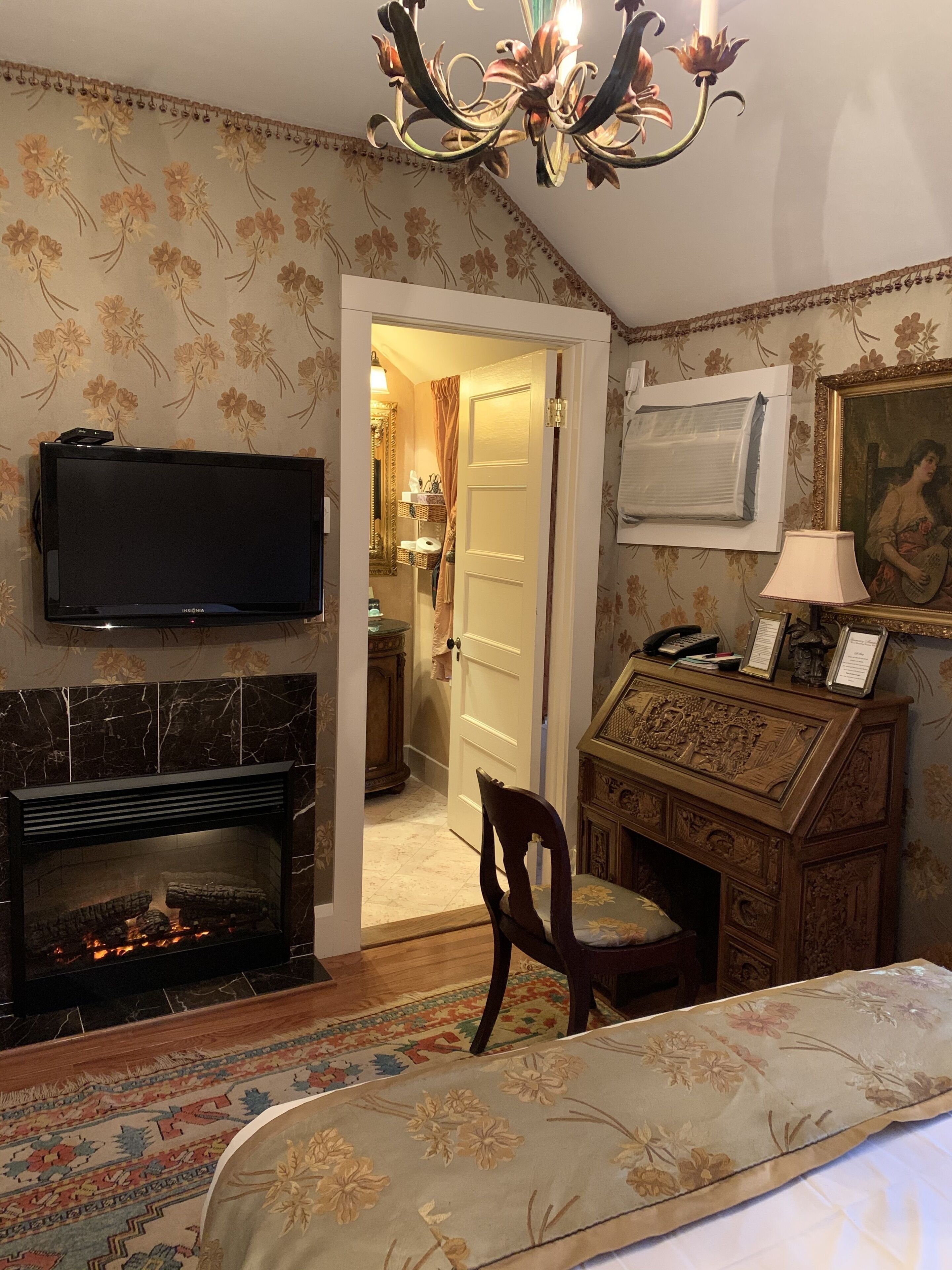 A room with a fireplace, a television, a desk with a lamp, and a door leading to another room.