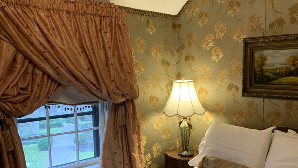 Un dormitorio con cama, mesita de noche con lámpara, papel tapiz floral y ventana con cortinas.