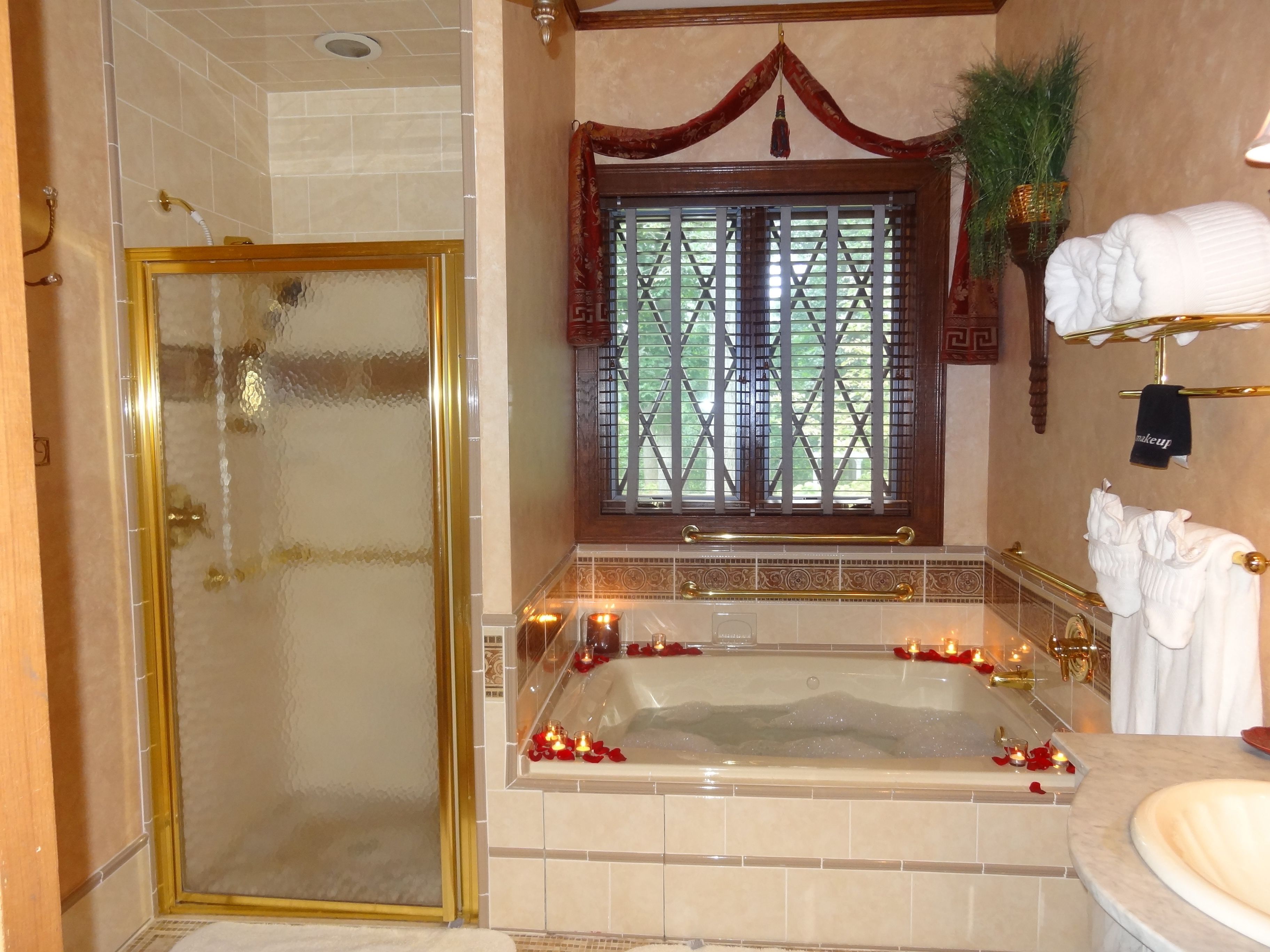 Une salle de bain dotée d’une douche à parois de verre, d’une baignoire sur pieds ornée de pétales de roses et d’une fenêtre agrémentée de rideaux décoratifs.