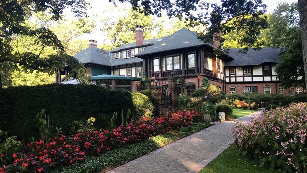 A traditional half-timbered house with a brick facade and a wooden balcony, surrounded by lush gardens and a paved walkway.