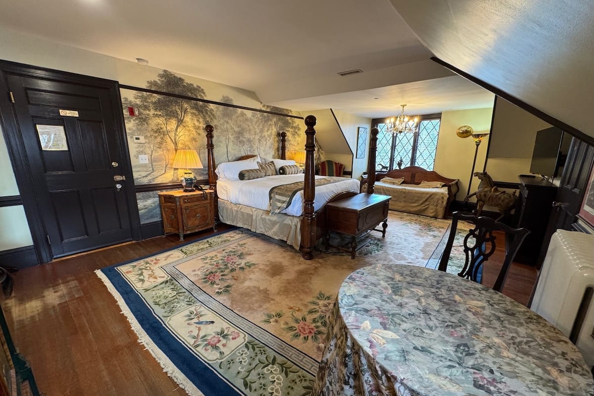 A bedroom with a four-poster bed, a nightstand, a dresser, and a chair. There is a large window with a view of trees and a chandelier.