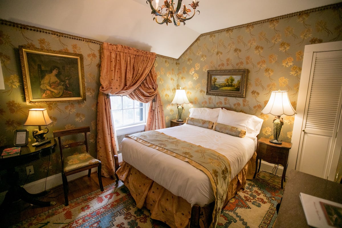 A bedroom with a large bed, two bedside lamps, a chair, a desk with a lamp, and floral wallpaper.