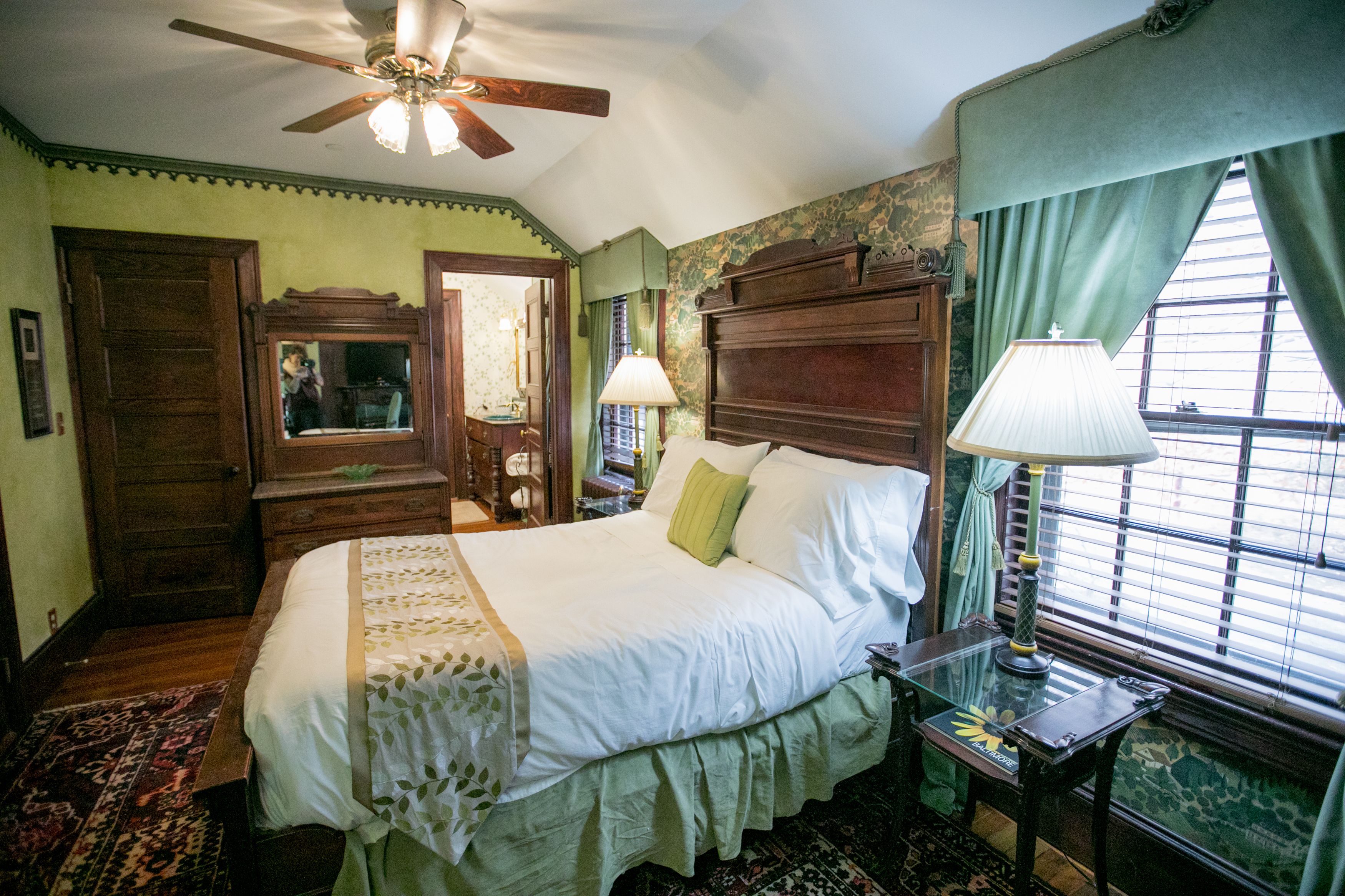 A traditional bedroom with a large bed, a ceiling fan, a window with curtains, and a wooden dresser.