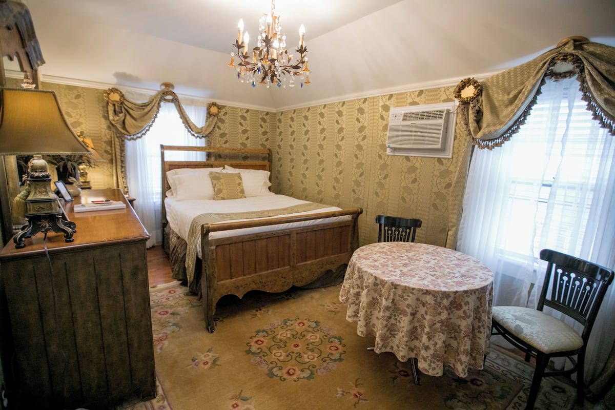 A bedroom with a wooden bed, a round table with a floral tablecloth, a chair, a chandelier, and a vintage lamp.