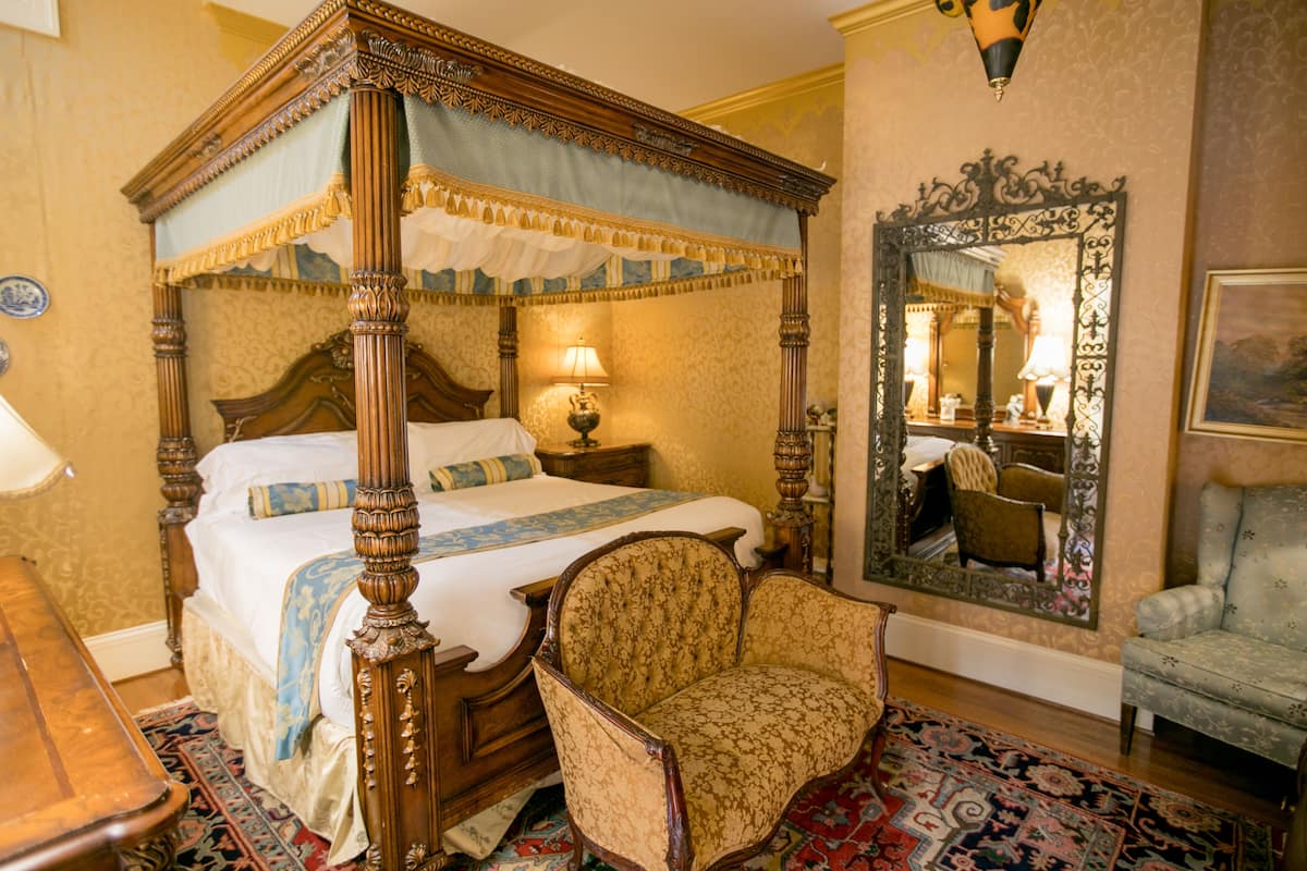A four-poster bed with a canopy, a wooden nightstand, a chair, a mirror, and a patterned rug.