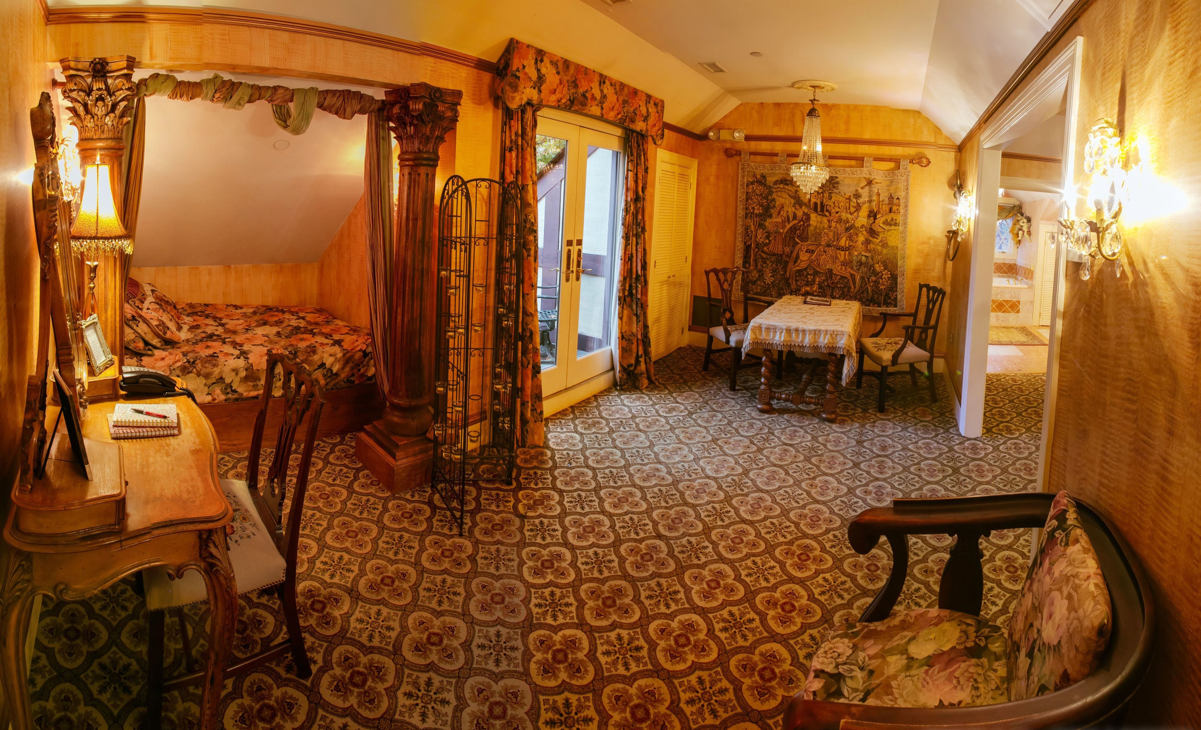 A room with a bed, a desk with a telephone, a chair, a table with a tablecloth, and a patterned floor.