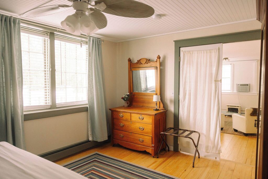 A bedroom with a wooden dresser, a mirror, a bed, a ceiling fan, and a view of a bathroom through an open door.