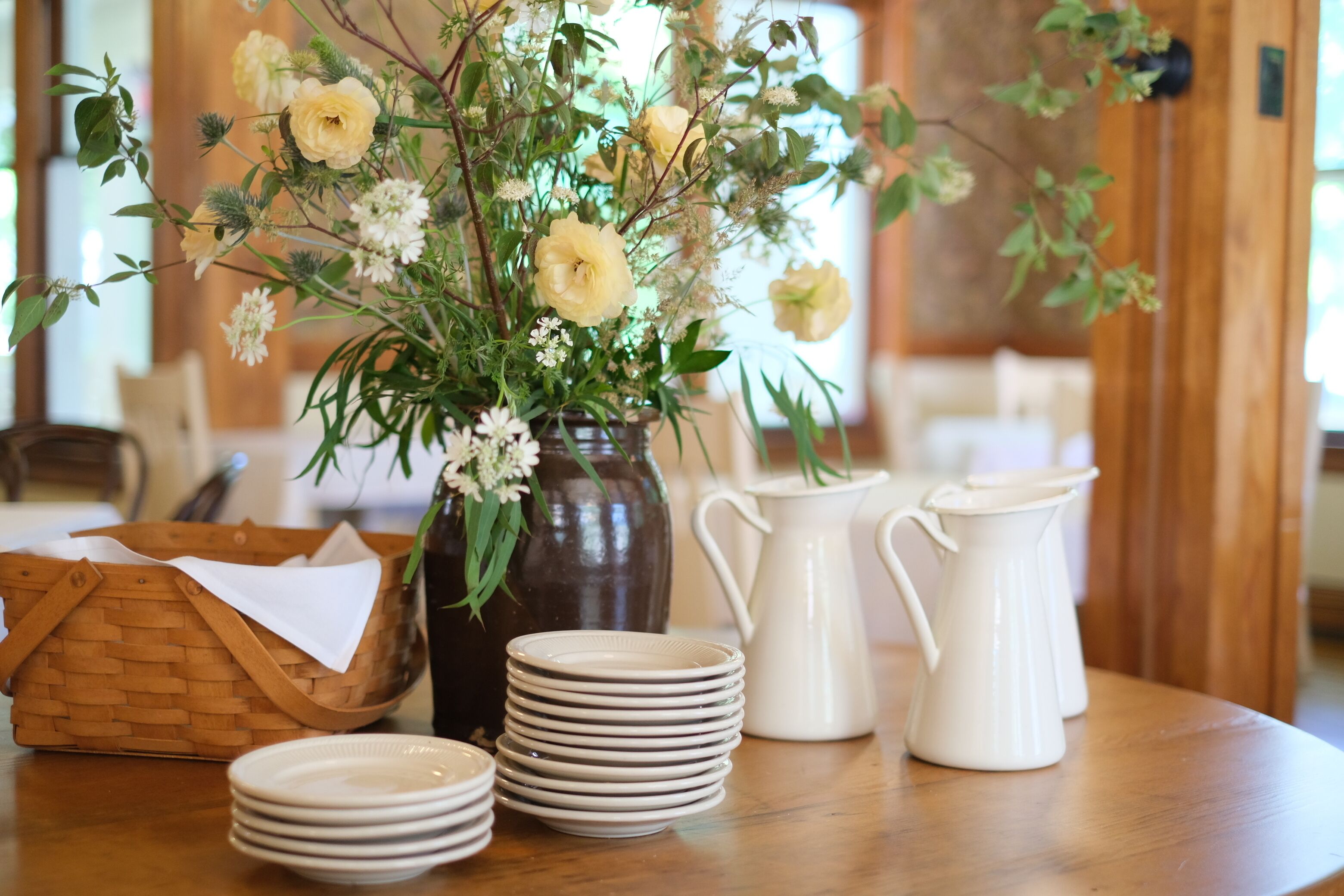 Une table avec un vase de fleurs, une pile d’assiettes, un pichet blanc et un panier en osier.