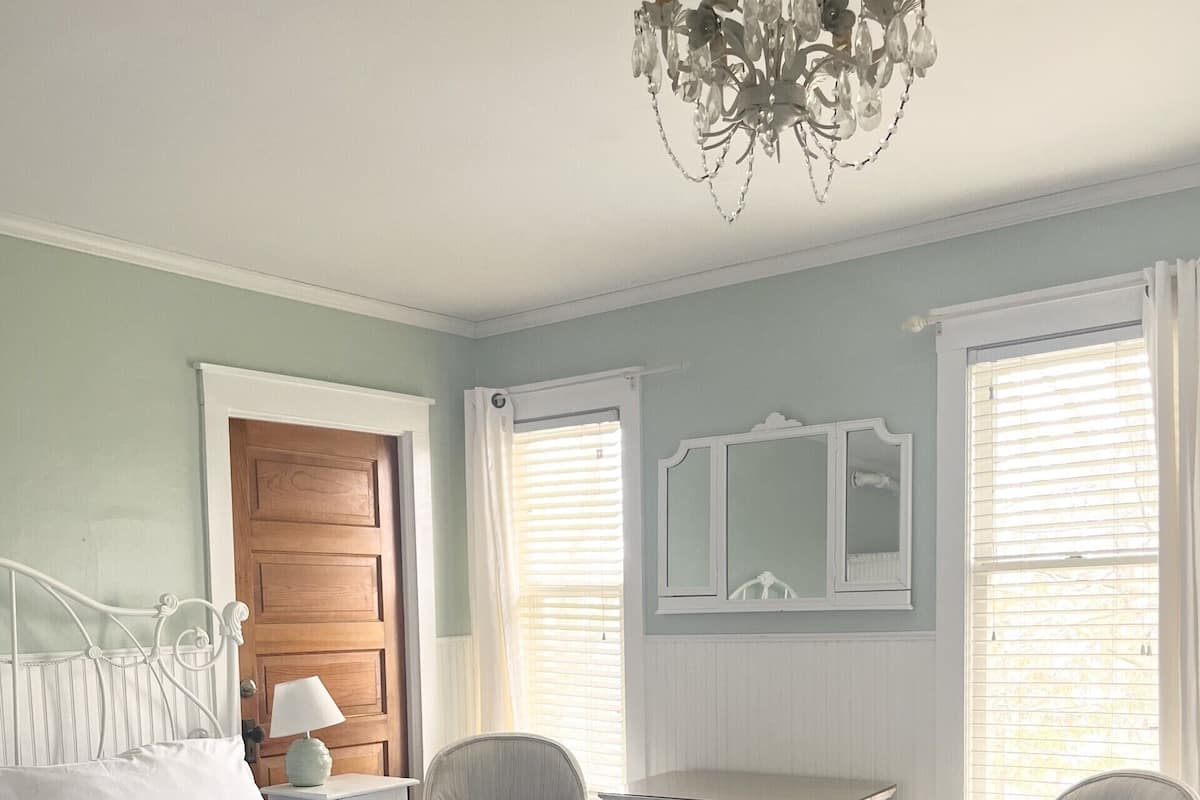 A bedroom with a bed, a desk, two chairs, a chandelier, and a window with blinds.