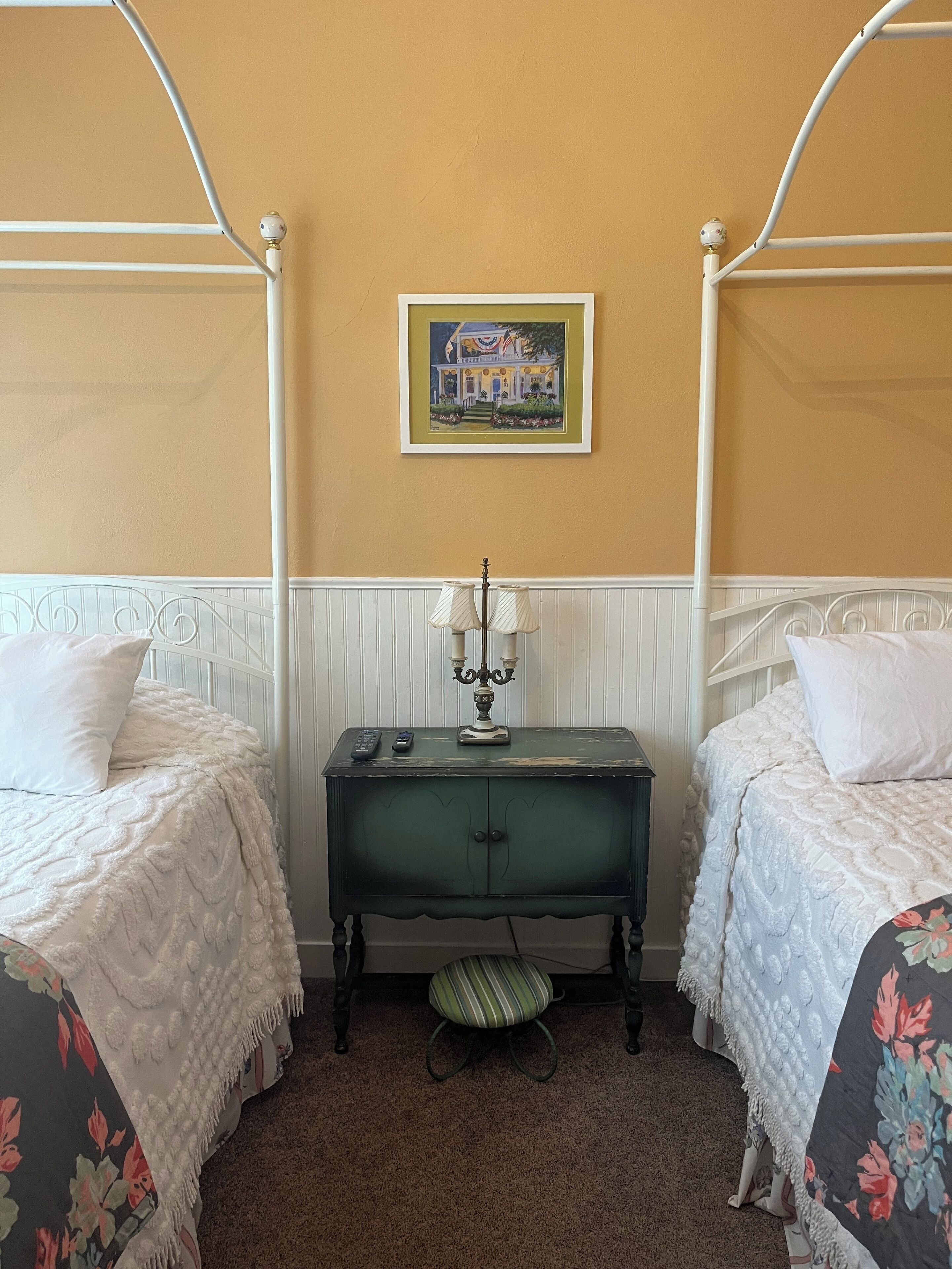 A bedroom with two single beds, a small table with a lamp, a framed picture on the wall, and a striped chair.