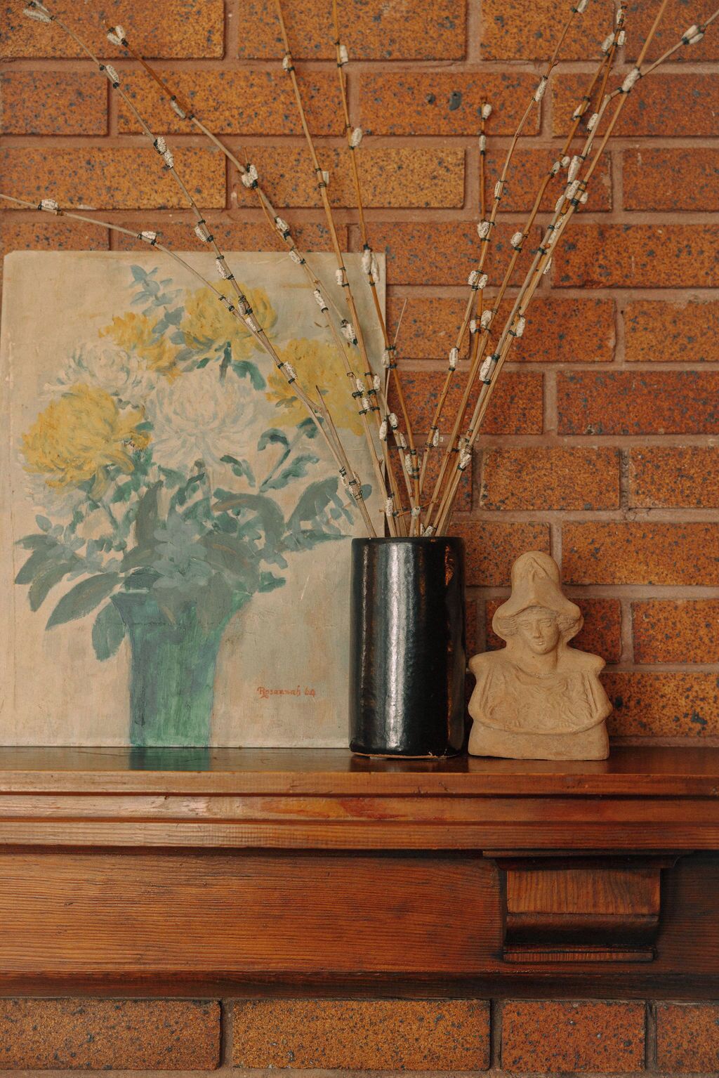 Et maleri af blomster i en vase, en metalkande og en skulptur på en træhylde foran en murstensvæg.