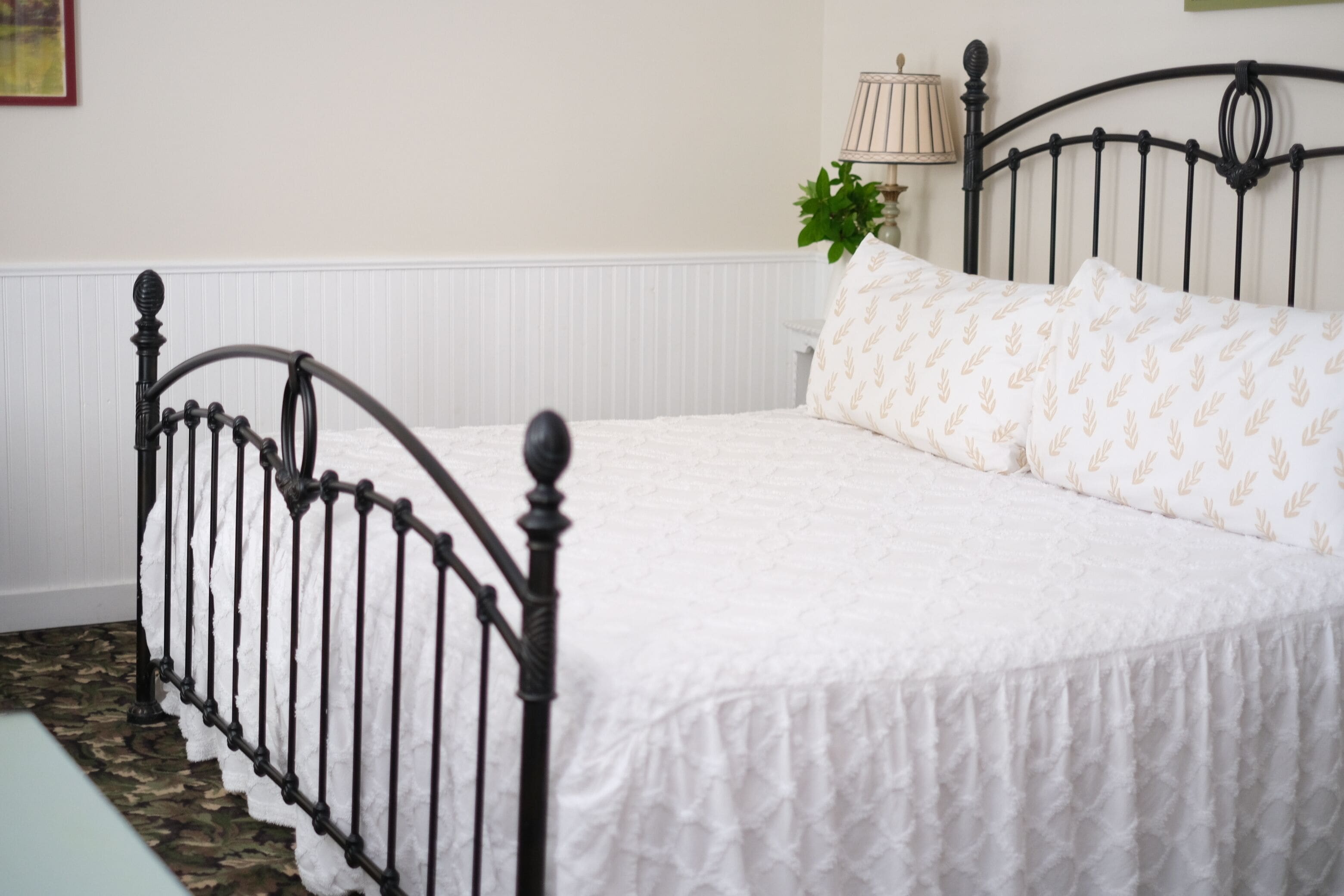 A neatly made bed with a white bedspread and matching pillows, a black metal headboard, and a green plant beside the bed.