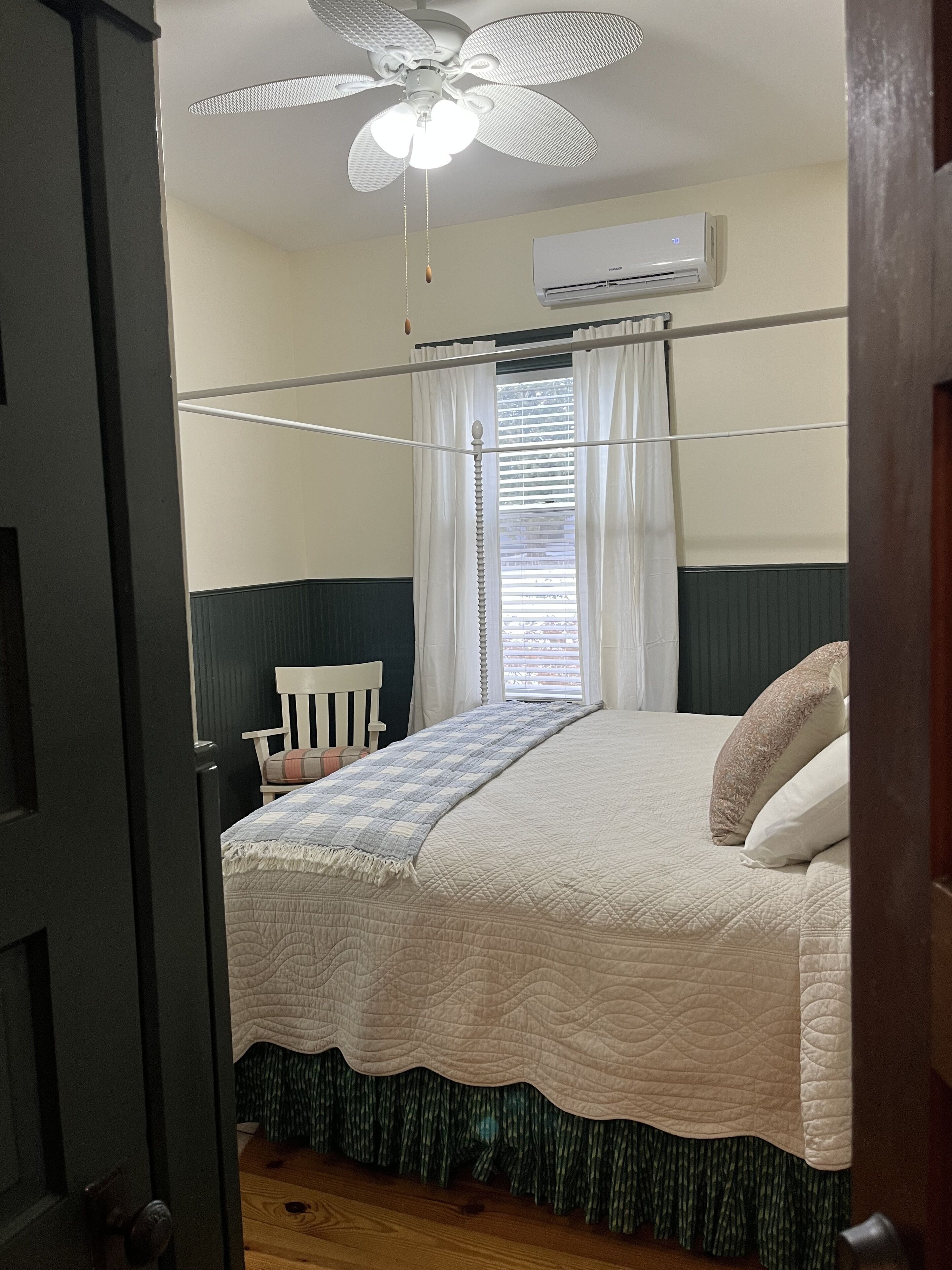 A bedroom with a bed, a ceiling fan, an air conditioning unit, a window with curtains, and a chair.