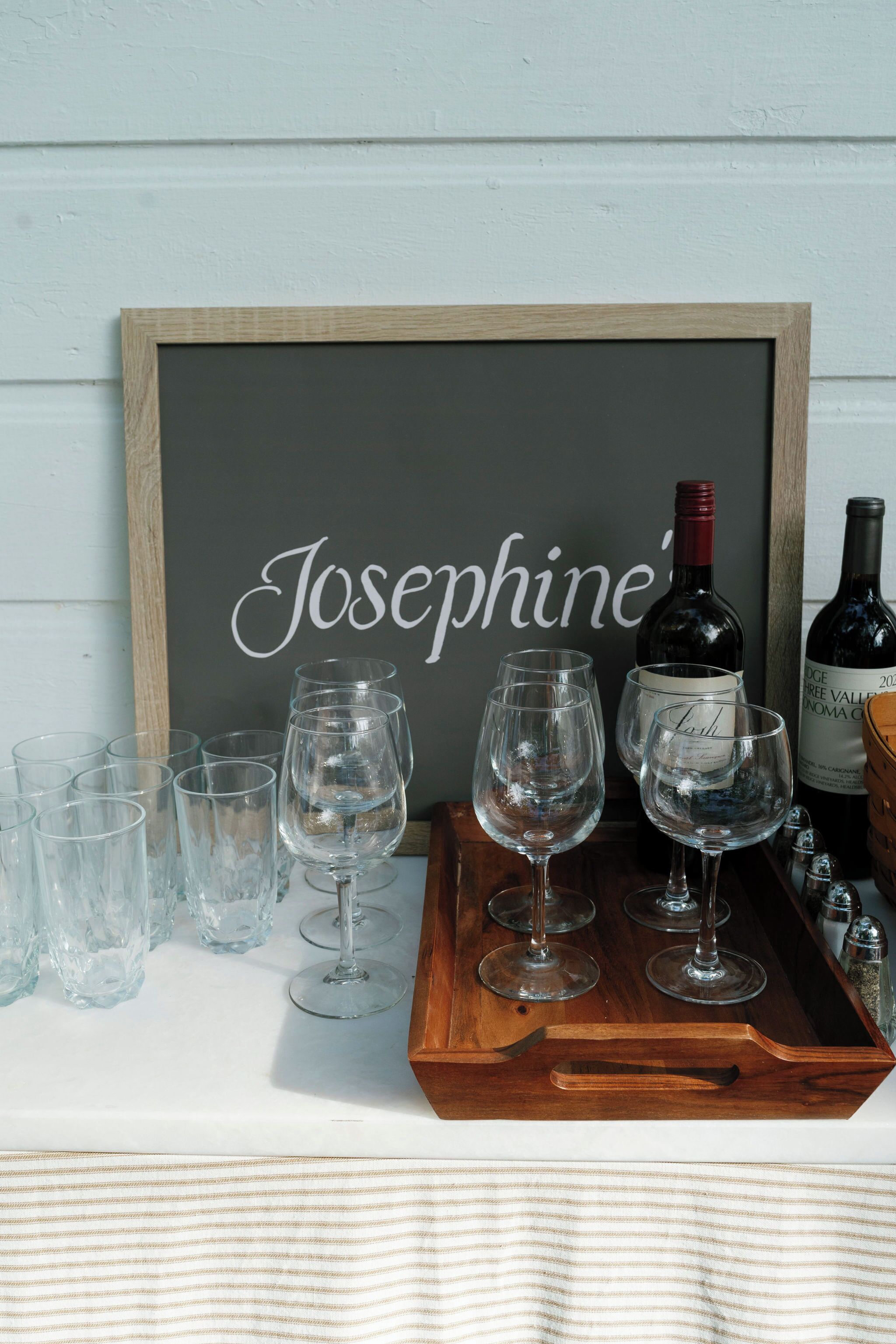 Ein Holztablett mit Weingläsern und Flaschen, ein Kreidetafel mit dem Namen 'Josephine' und ein gestreifter Stoff am unteren Rand.
