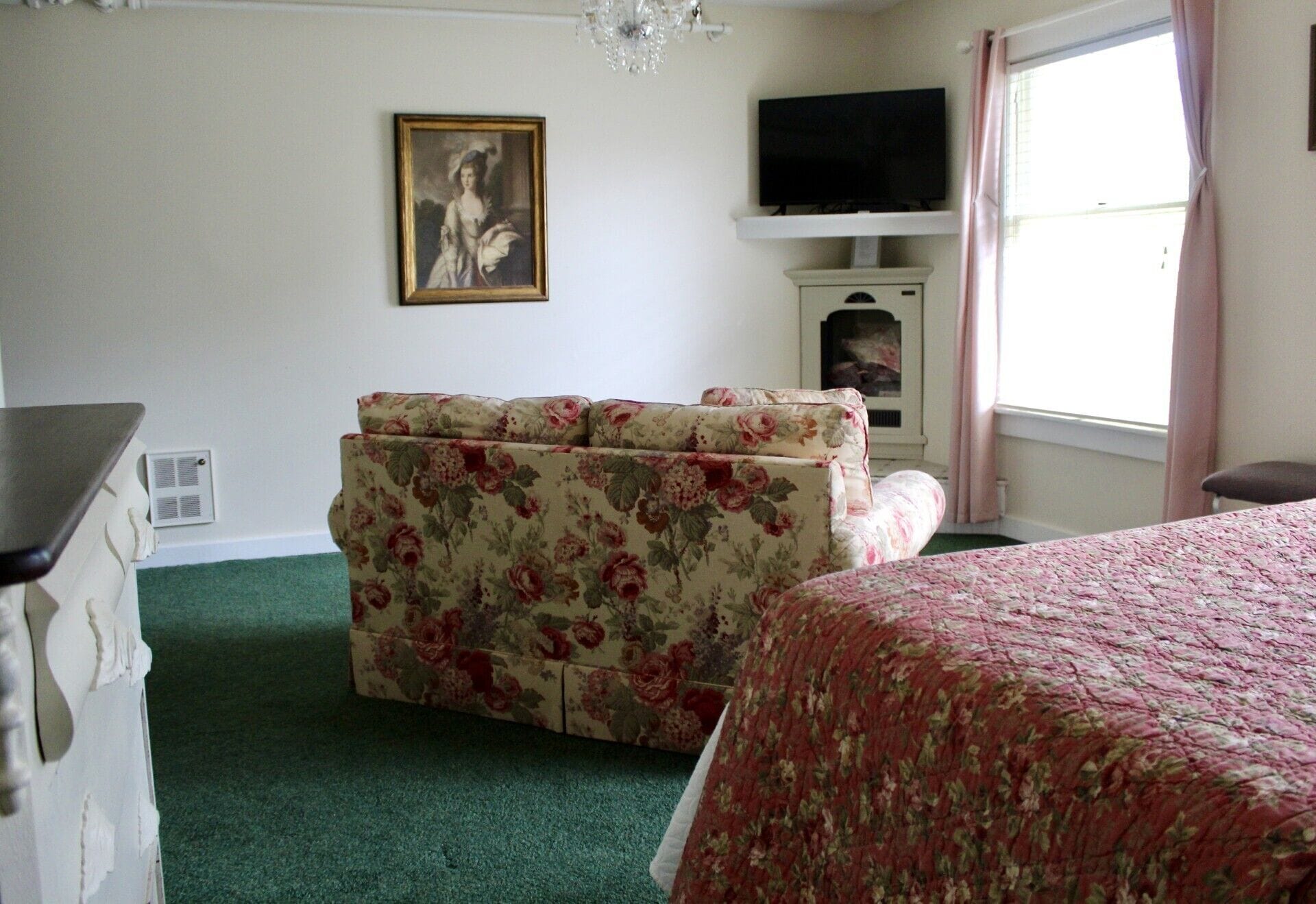 A bedroom with a floral sofa, a bed with a floral bedspread, a television on a shelf, and a painting on the wall.