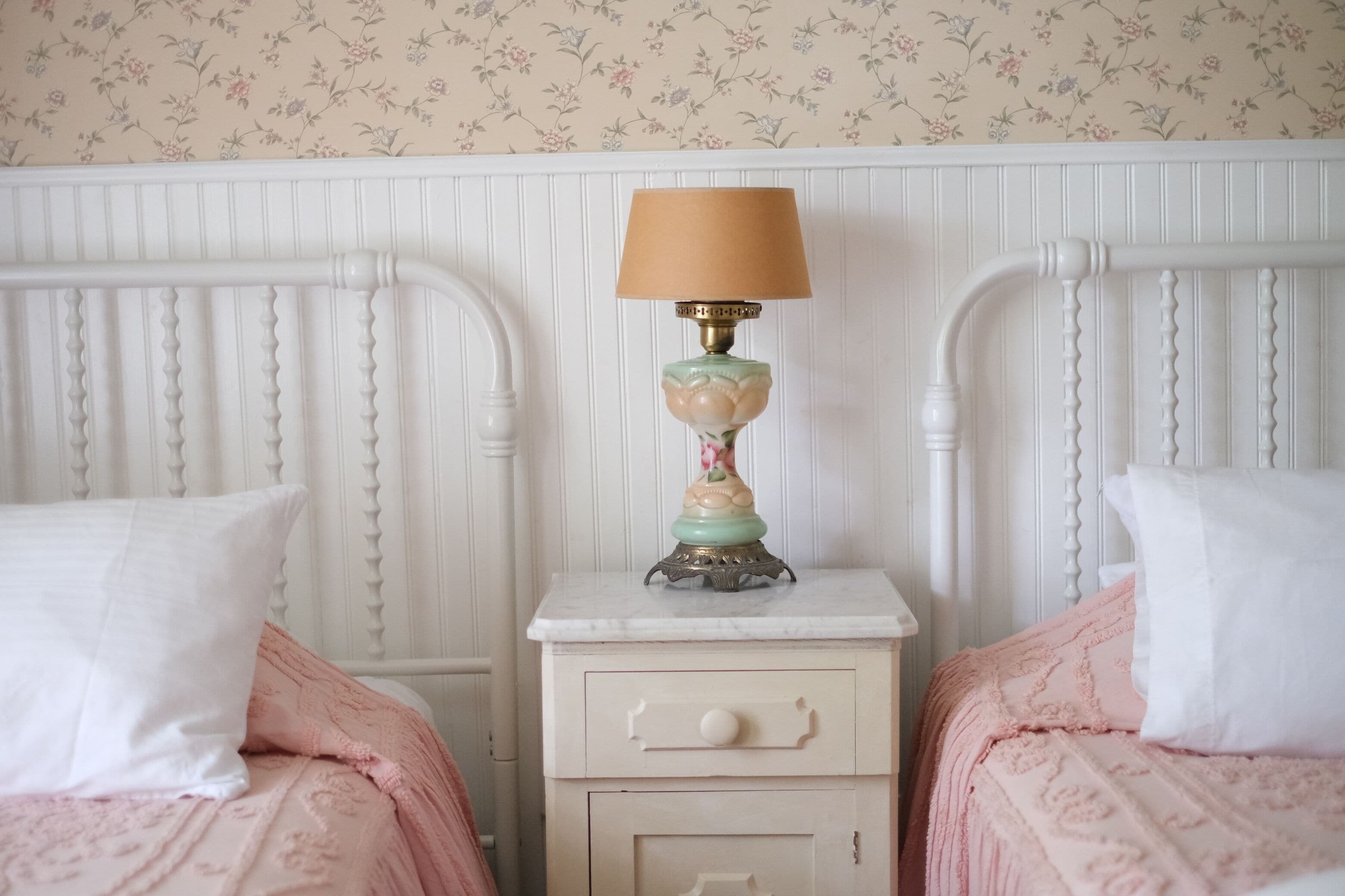 A bedroom with two beds, a nightstand with a lamp, and a floral wallpaper pattern.