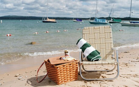 A beach chair with a striped cushion and a wicker basket with a bottle on the sand near the water.