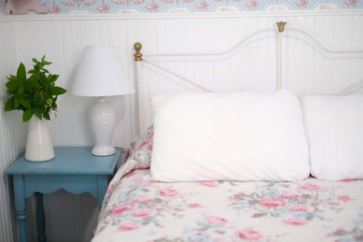 A bedroom with a floral bedspread, a white lamp, and a small plant on a blue nightstand.