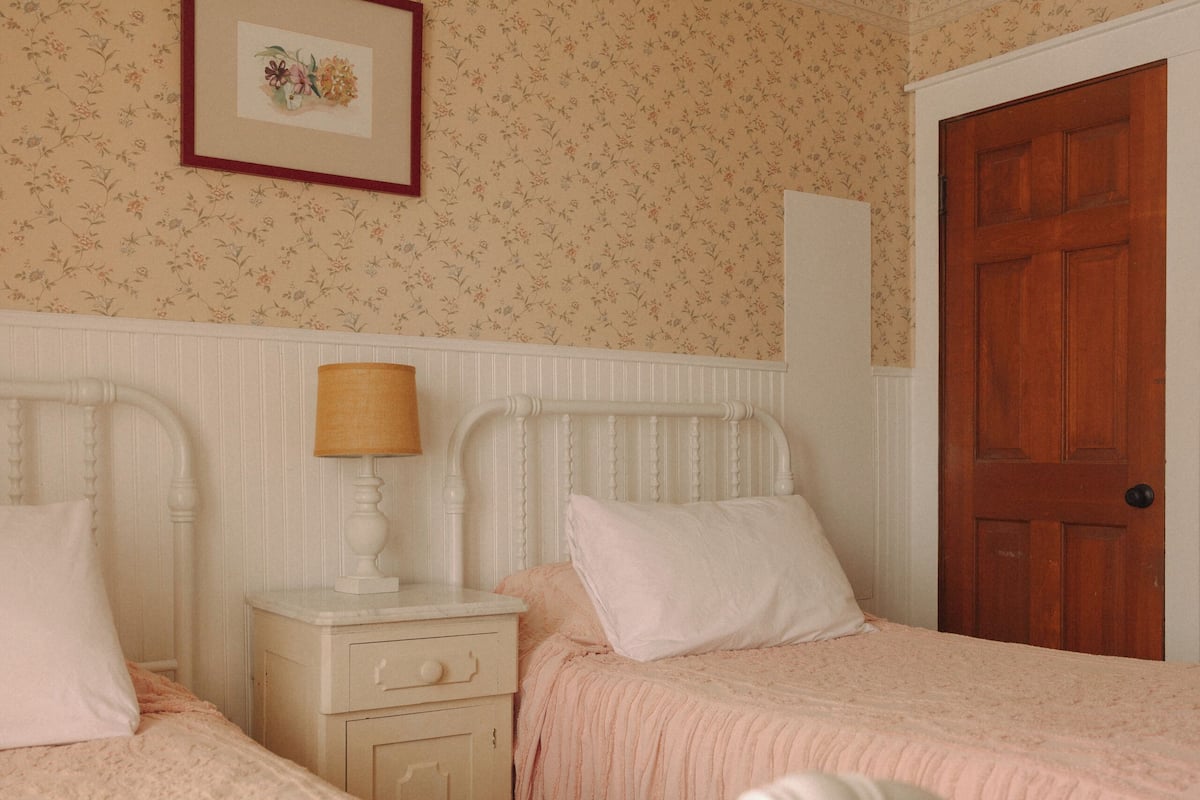 A bedroom with two beds, a nightstand with a lamp, a framed picture on the wall, and a wooden door.