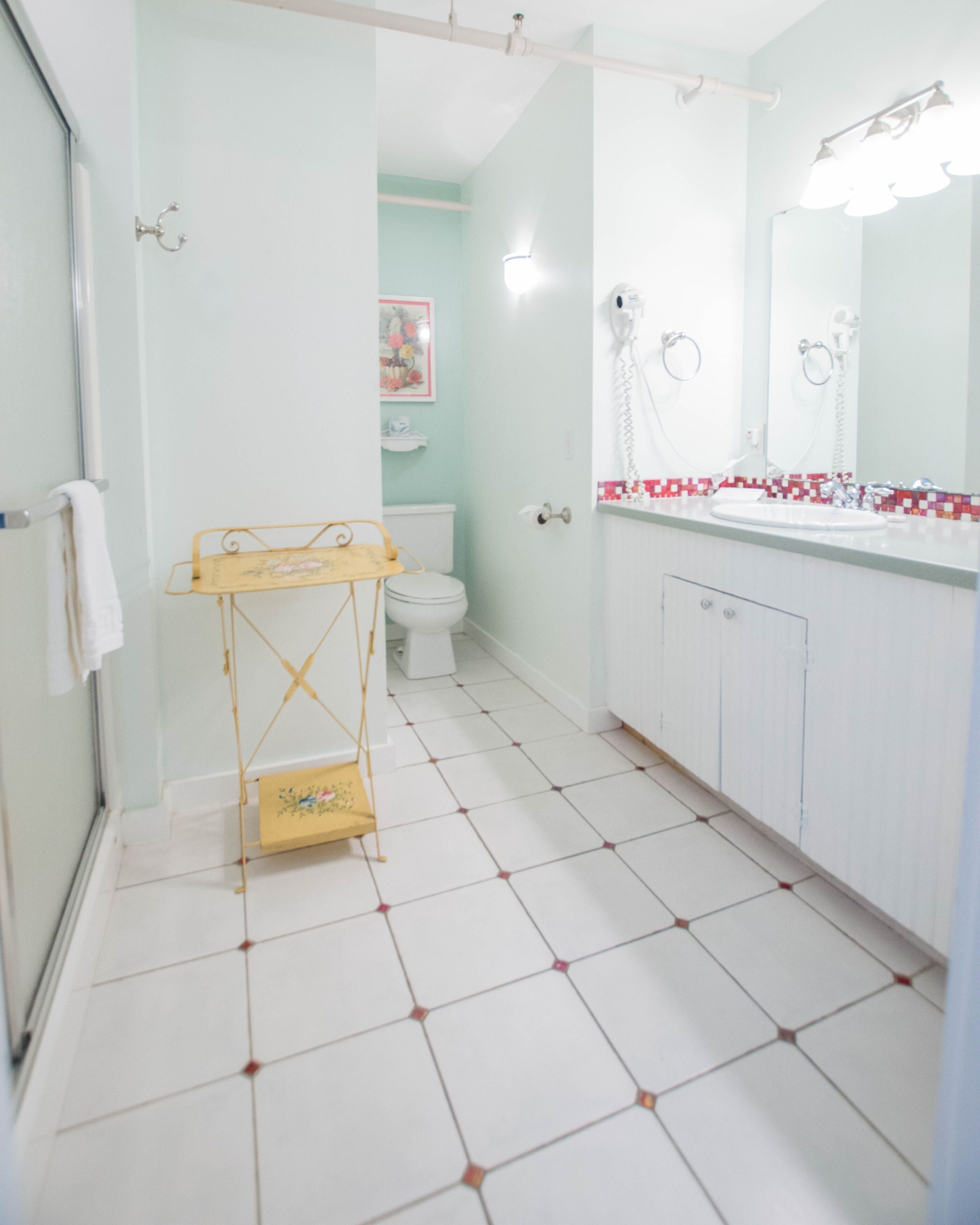 Un baño con un inodoro blanco, un tendedero amarillo, un lavabo blanco con espejo y suelo de baldosas.
